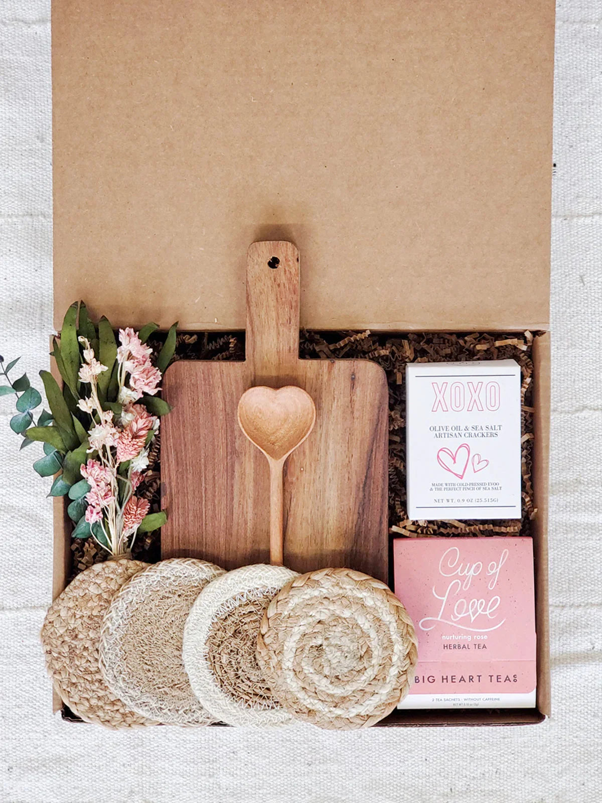 Gift box with wooden cutting board, heart-shaped wooden spoon, artisan crackers, herbal tea, dried flowers, and woven coasters