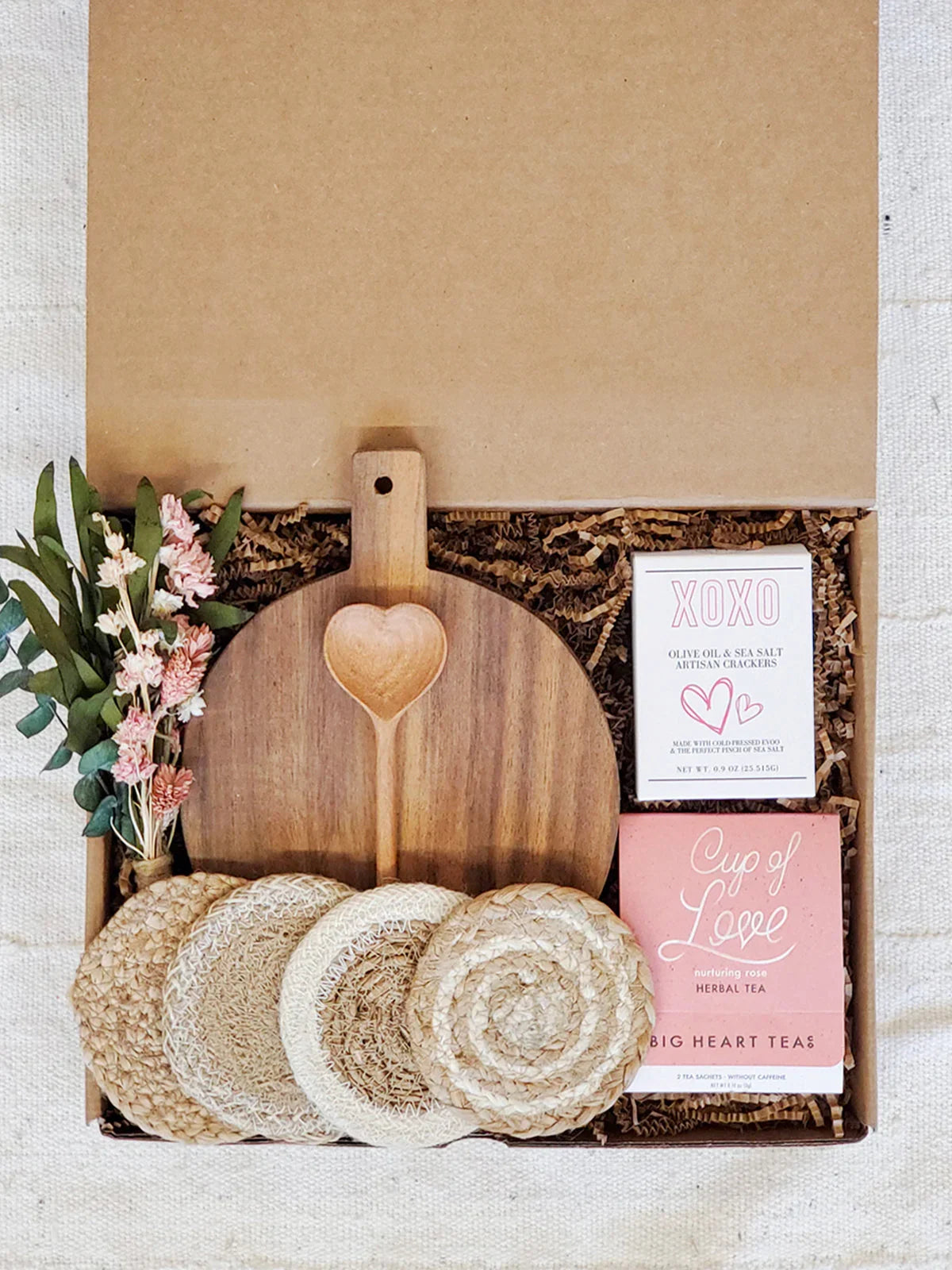 Gift box with round wooden cutting board, heart-shaped spoon, coasters, herbal tea, crackers, and dried flowers