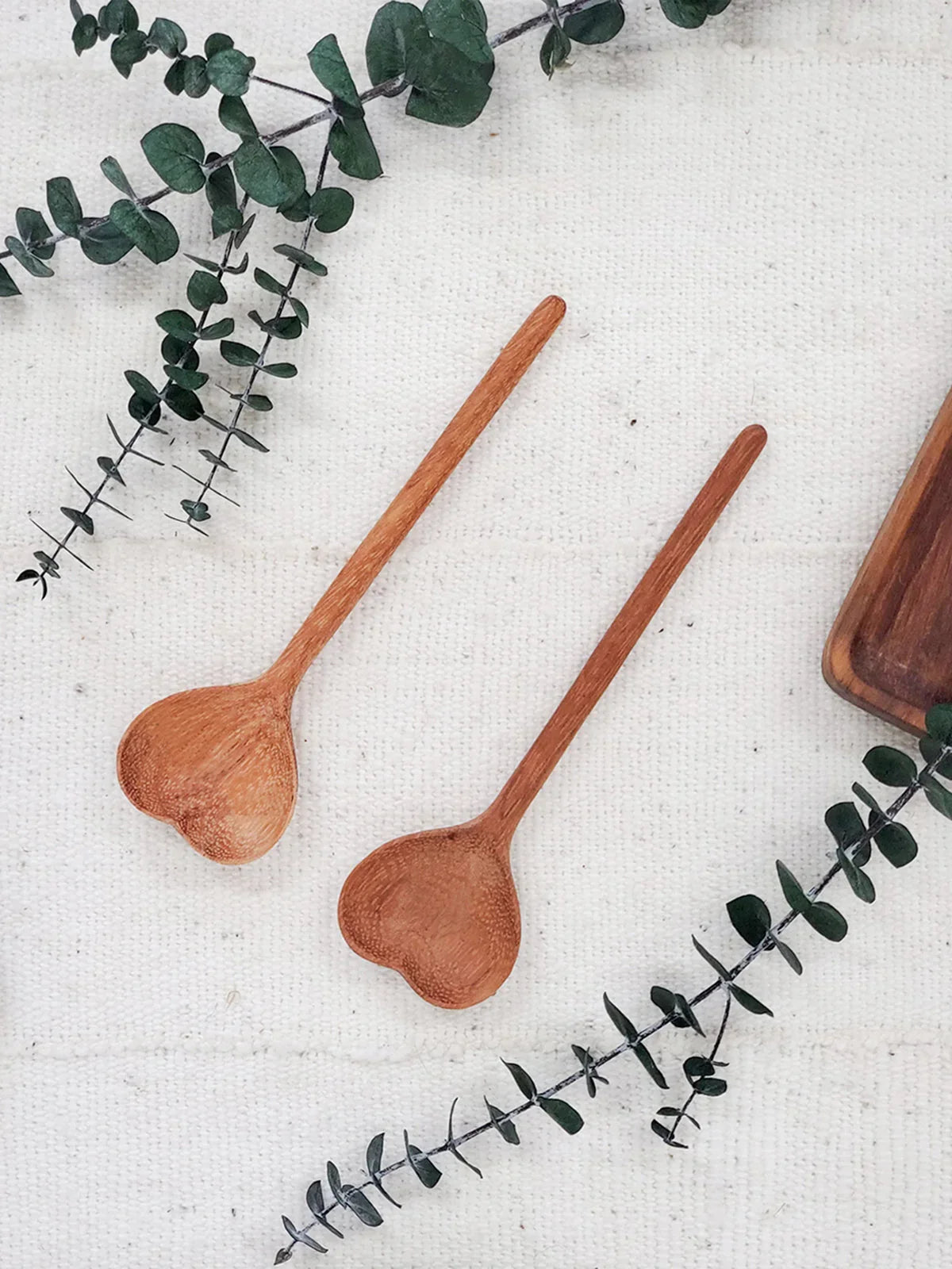 Two wooden heart-shaped spoons on white fabric with green eucalyptus branches and a wooden tray corner