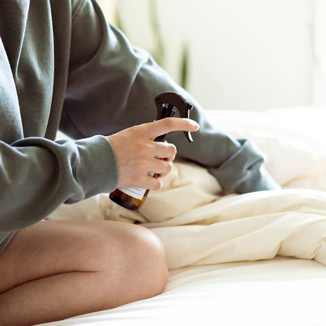 Person in cozy loungewear spraying eco-friendly mist on cream bedding, Qu!rk Boutique