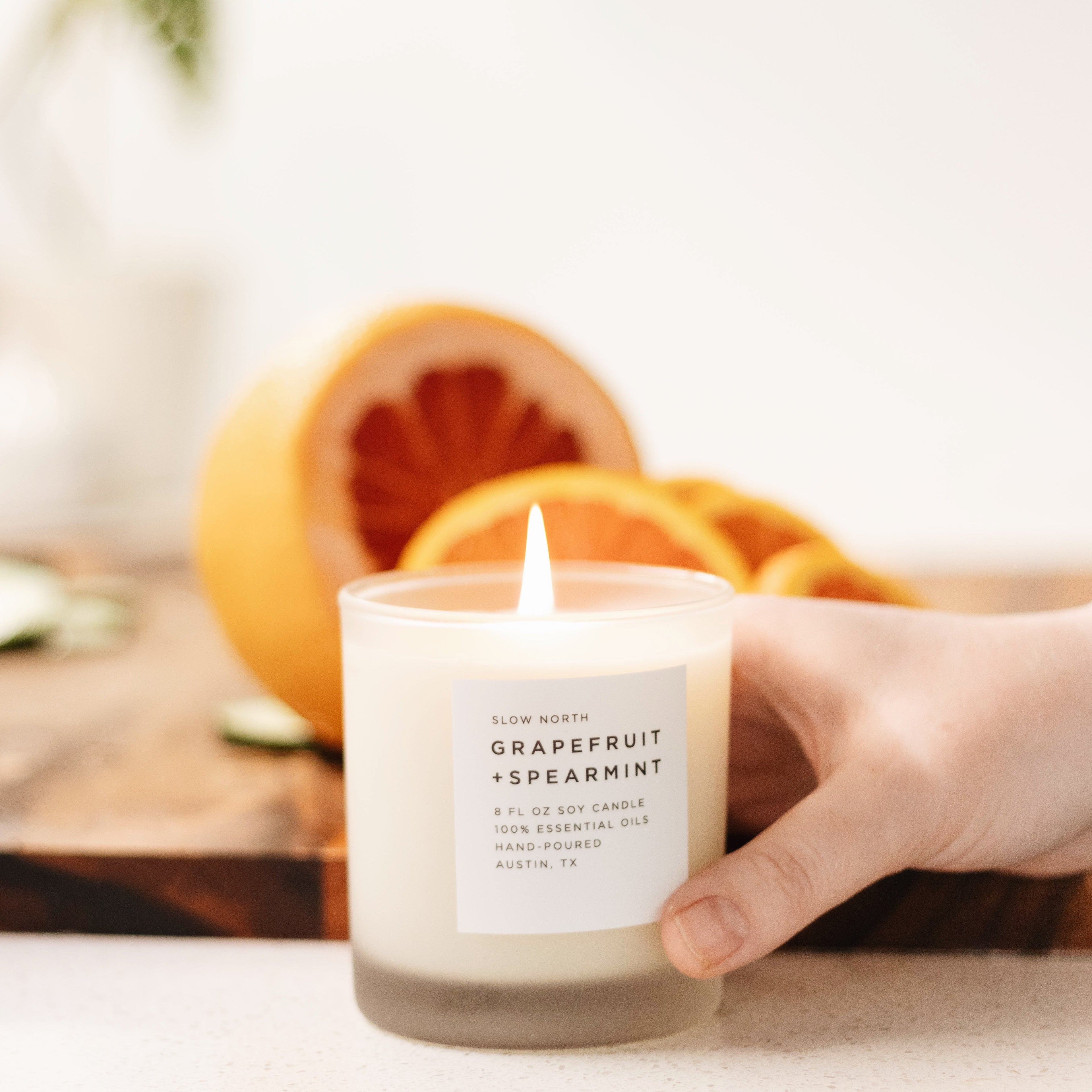 Hand holding lit Slow North Grapefruit + Spearmint soy candle with sliced grapefruit in background