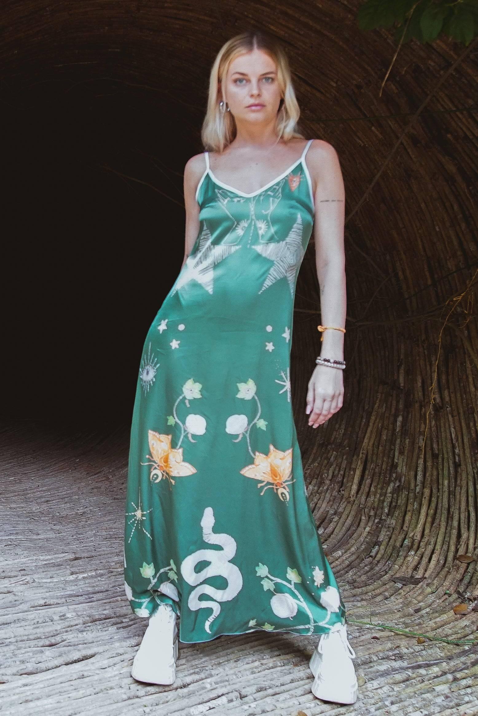 Blonde woman wearing a green satin maxi dress with floral and snake prints, standing in woven tunnel
