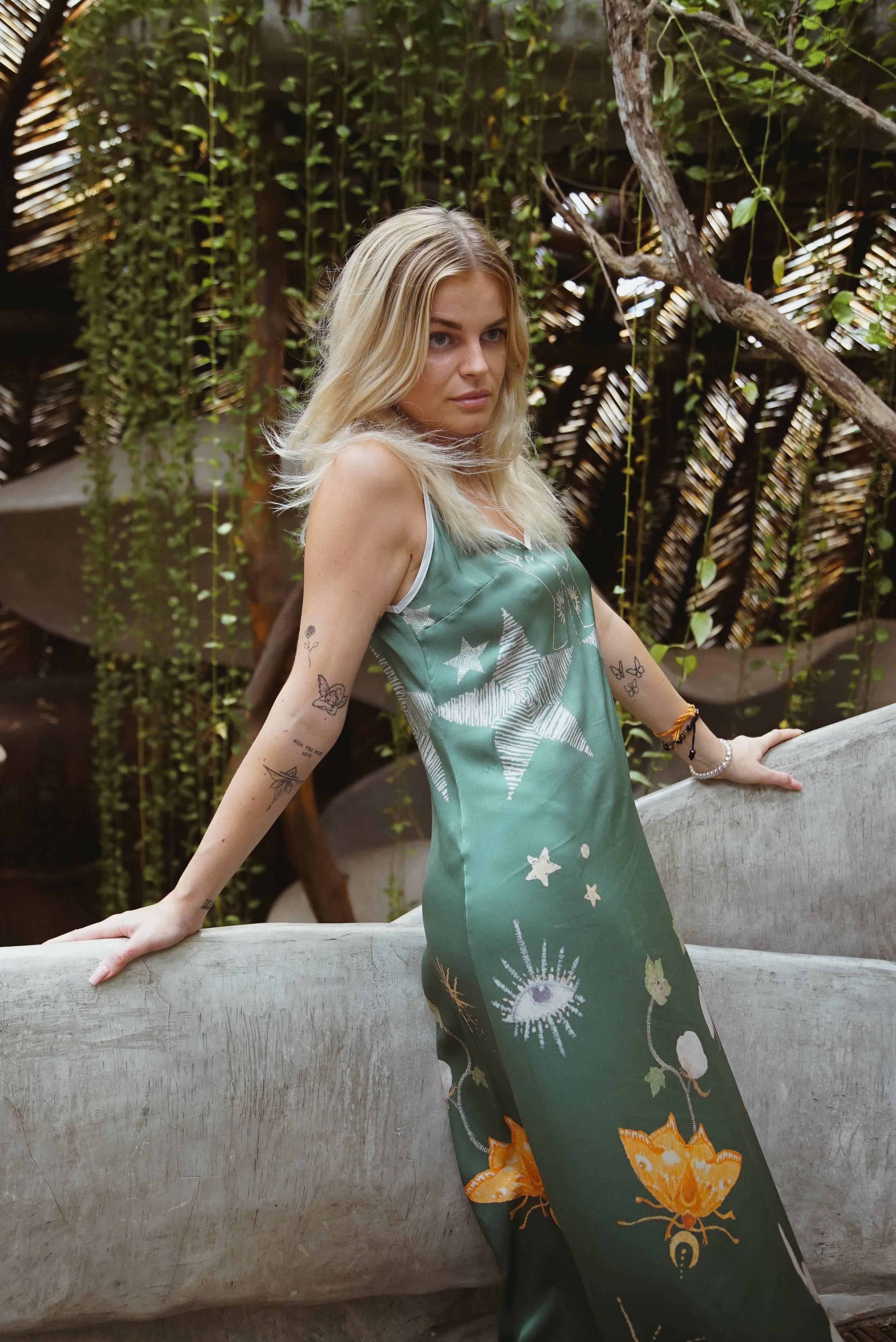 Blonde woman in a green satin maxi dress with star and eye prints, posing outdoors by concrete railing