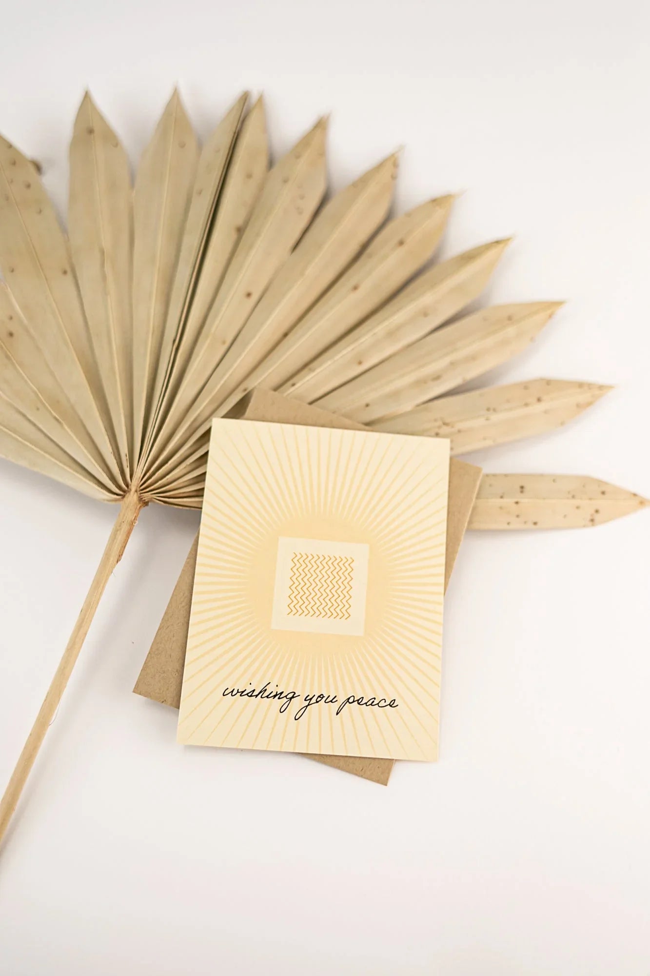 Greeting card with 'wishing you peace' text on beige sunburst design, next to dried palm leaf