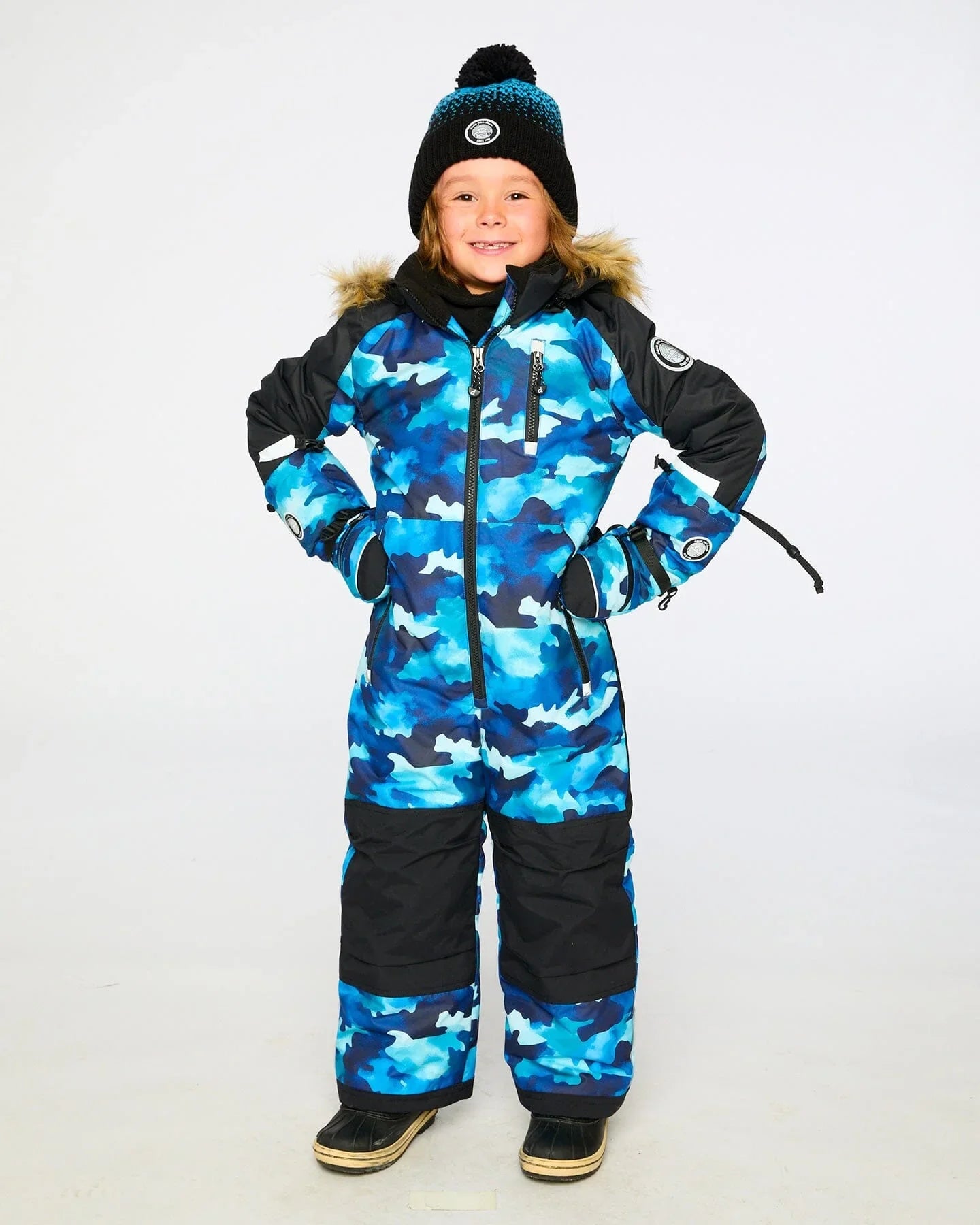 Smiling child in a blue camo snowsuit and winter hat, Qu!rk Boutique winter fashion.