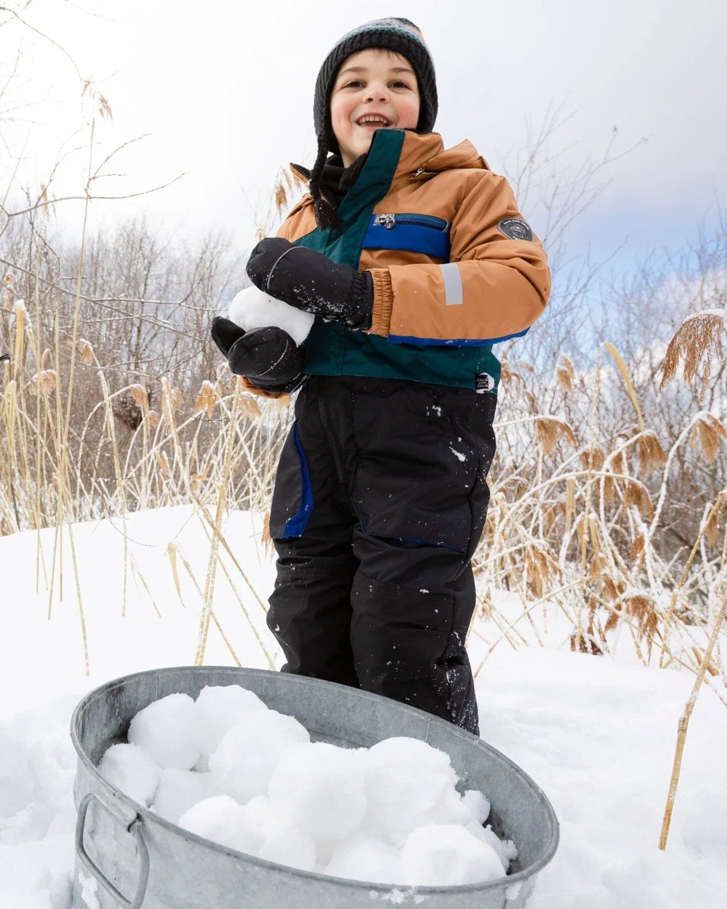 Smiling child in winter jacket and hat holding snowball outdoors by a bucket of snowballs