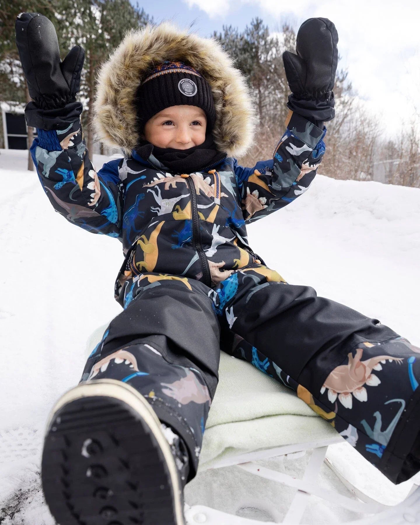 Smiling child in dinosaur snowsuit and fur hood, sledding on snow at Qu!rk Boutique winter collection