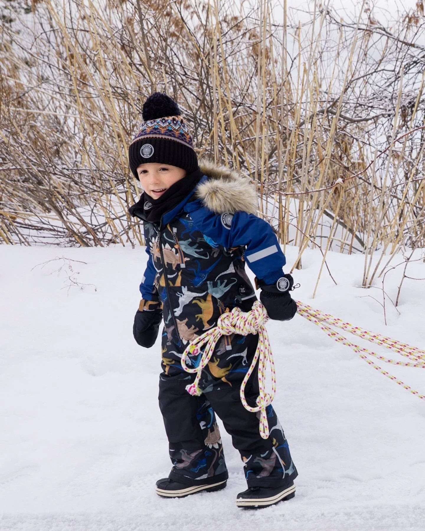 Child in colorful snowsuit and knit hat holding rope, playing in snowy winter landscape