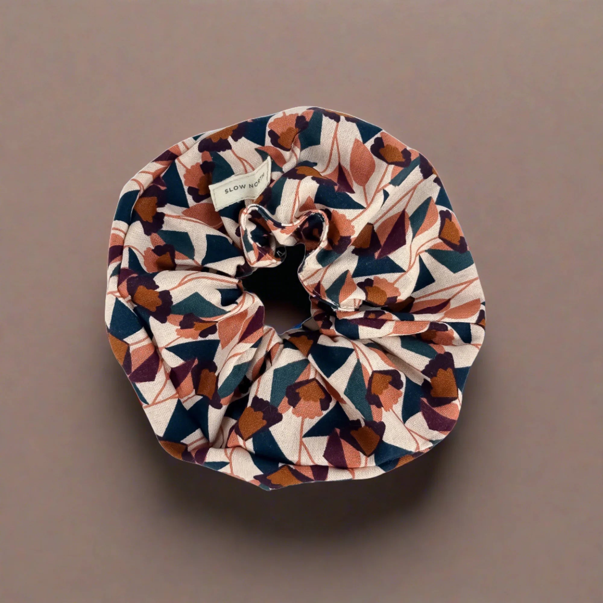 Floral patterned fabric scrunchie with earthy tones on a brown background, labeled Slow North
