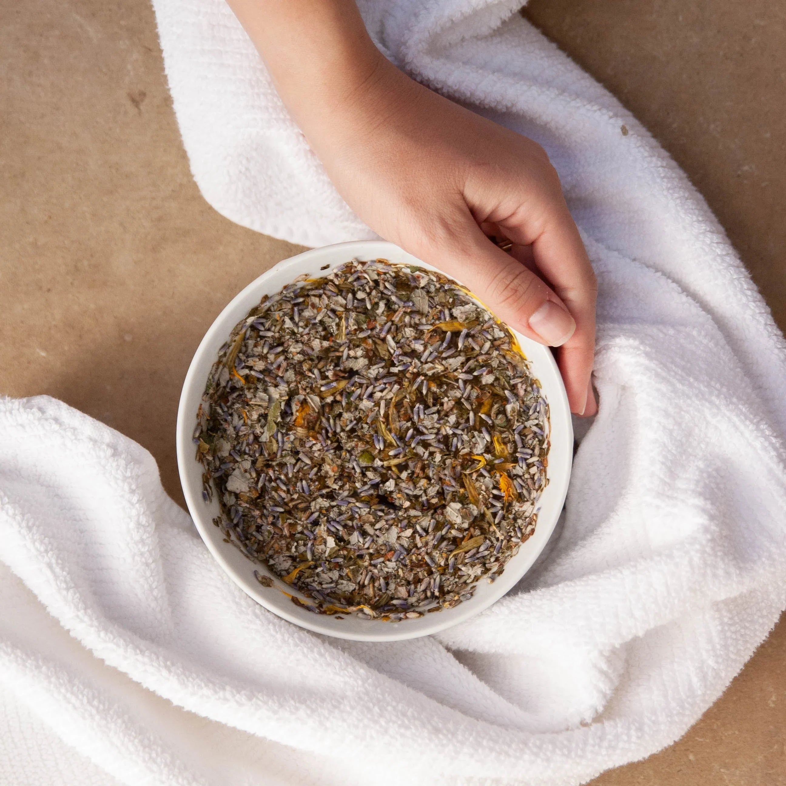 Hand holding bowl of herbal facial steam blend on white towel, Qu!rk Boutique skincare product