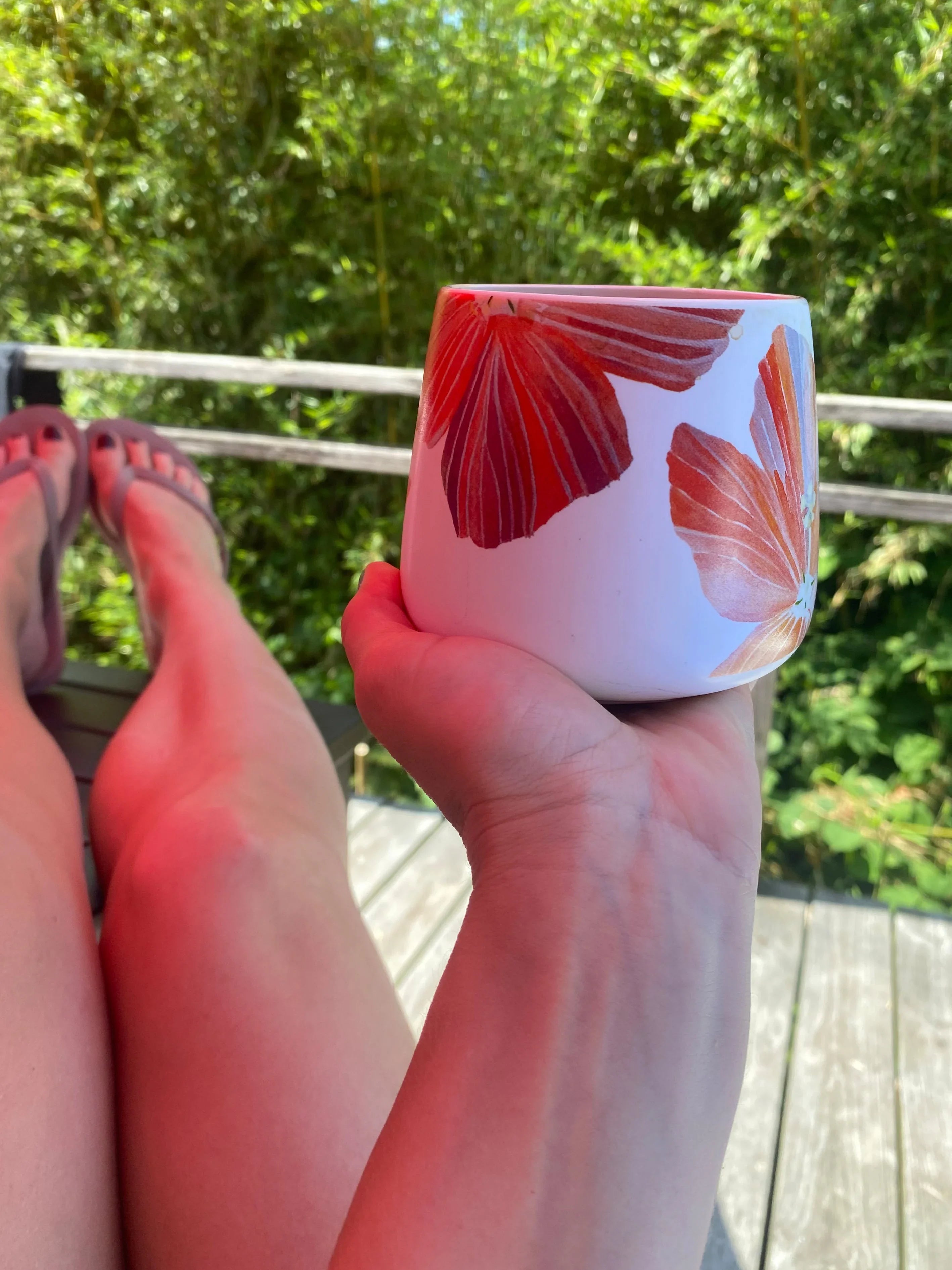 Person holding a white mug with red hibiscus flowers while relaxing on a wooden deck surrounded by green foliage