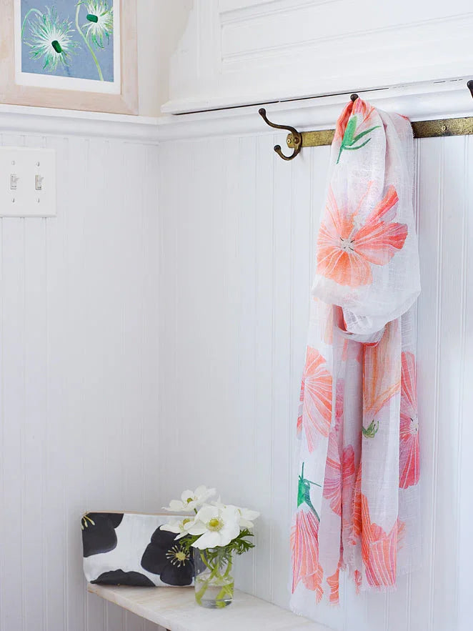 White wall with brass coat rack holding floral scarf, framed flower art, white flowers in vase on marble shelf