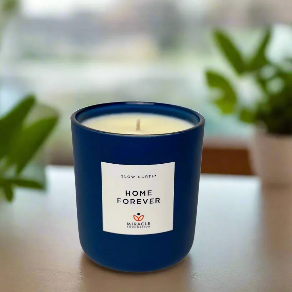 Slow North Home Forever candle in a blue glass jar on a table, with plants in the background at Qu!rk Boutique.