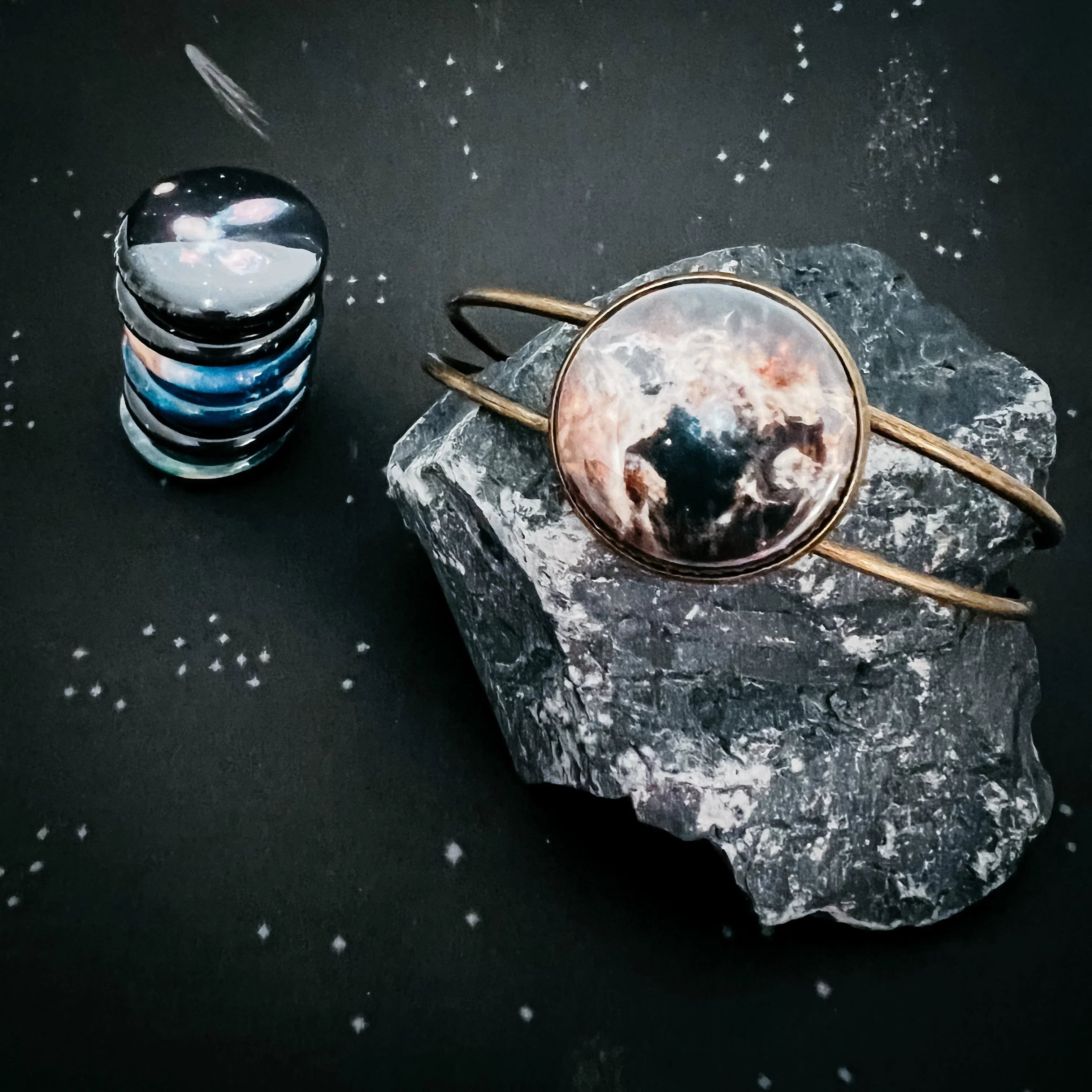Round cosmic-themed bracelet on dark textured rock and stacked planet-like round stones on black starry background