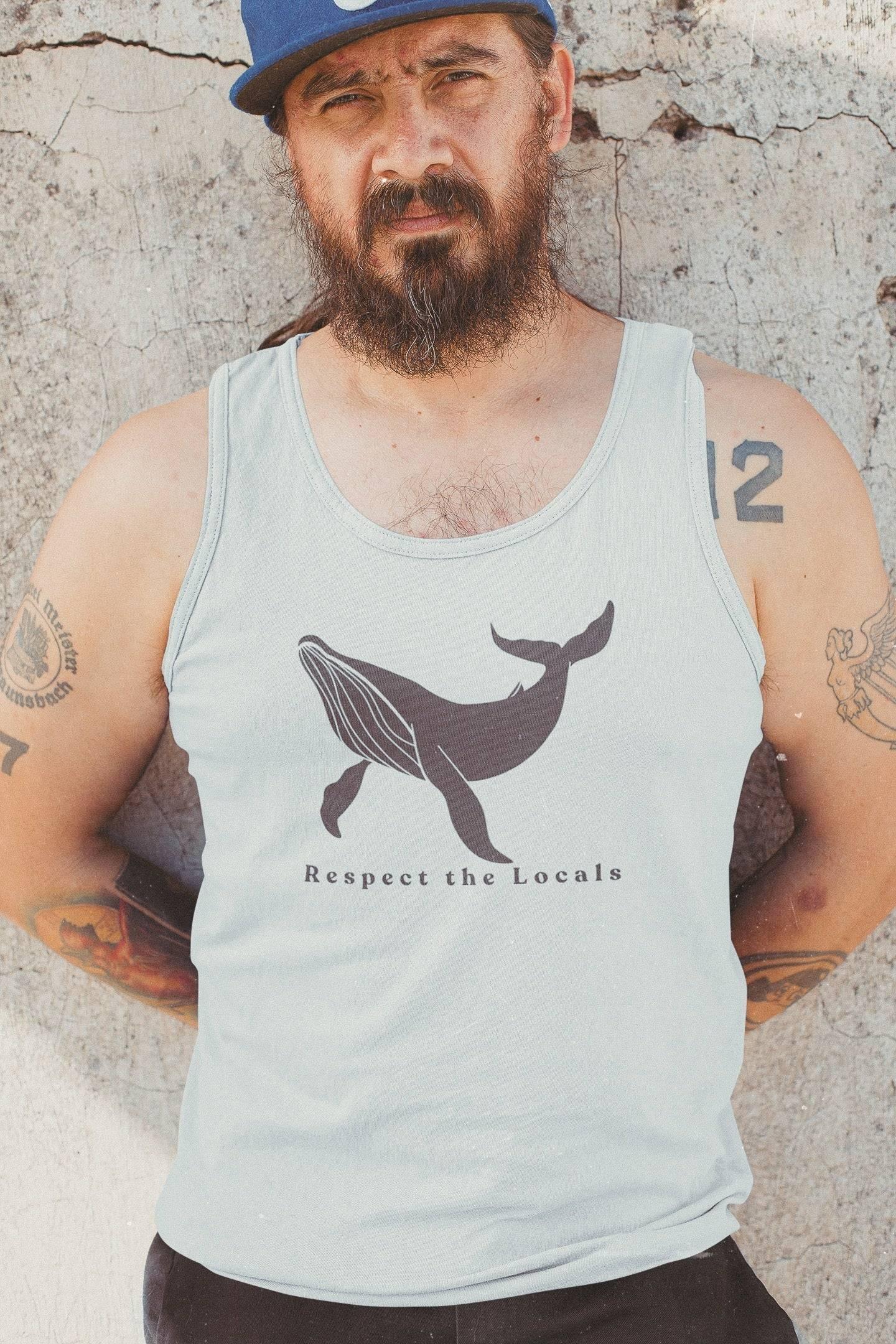 Bearded man wearing a light tank top with a whale graphic and the text Respect the Locals, standing against a cracked wall