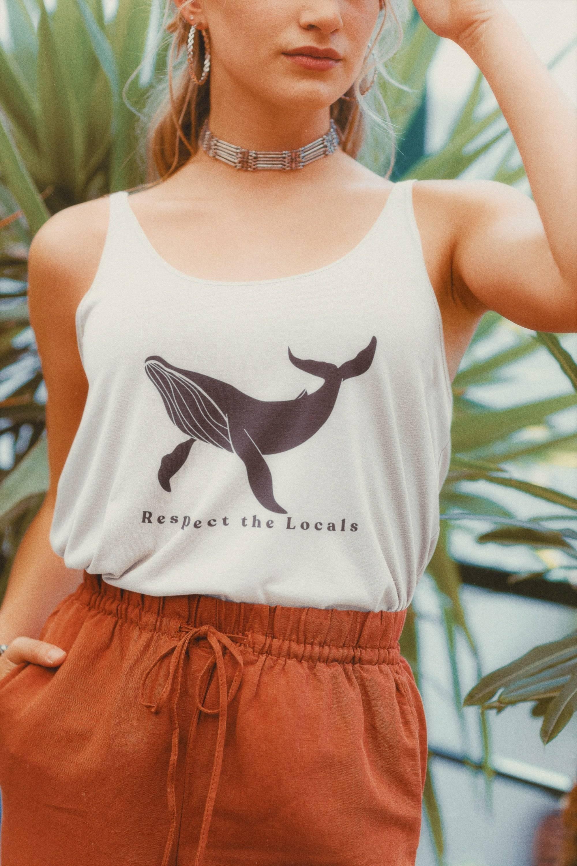 Close-up of woman wearing white tank top with whale graphic and text 'Respect the Locals', rust-colored pants