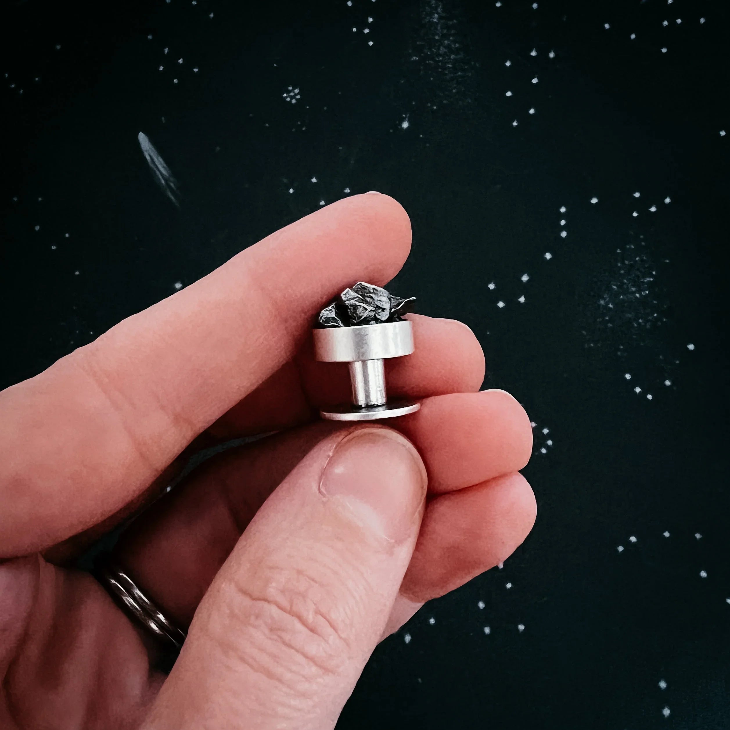 Hand holding a silver stud earring with a rough black stone against a dark speckled background