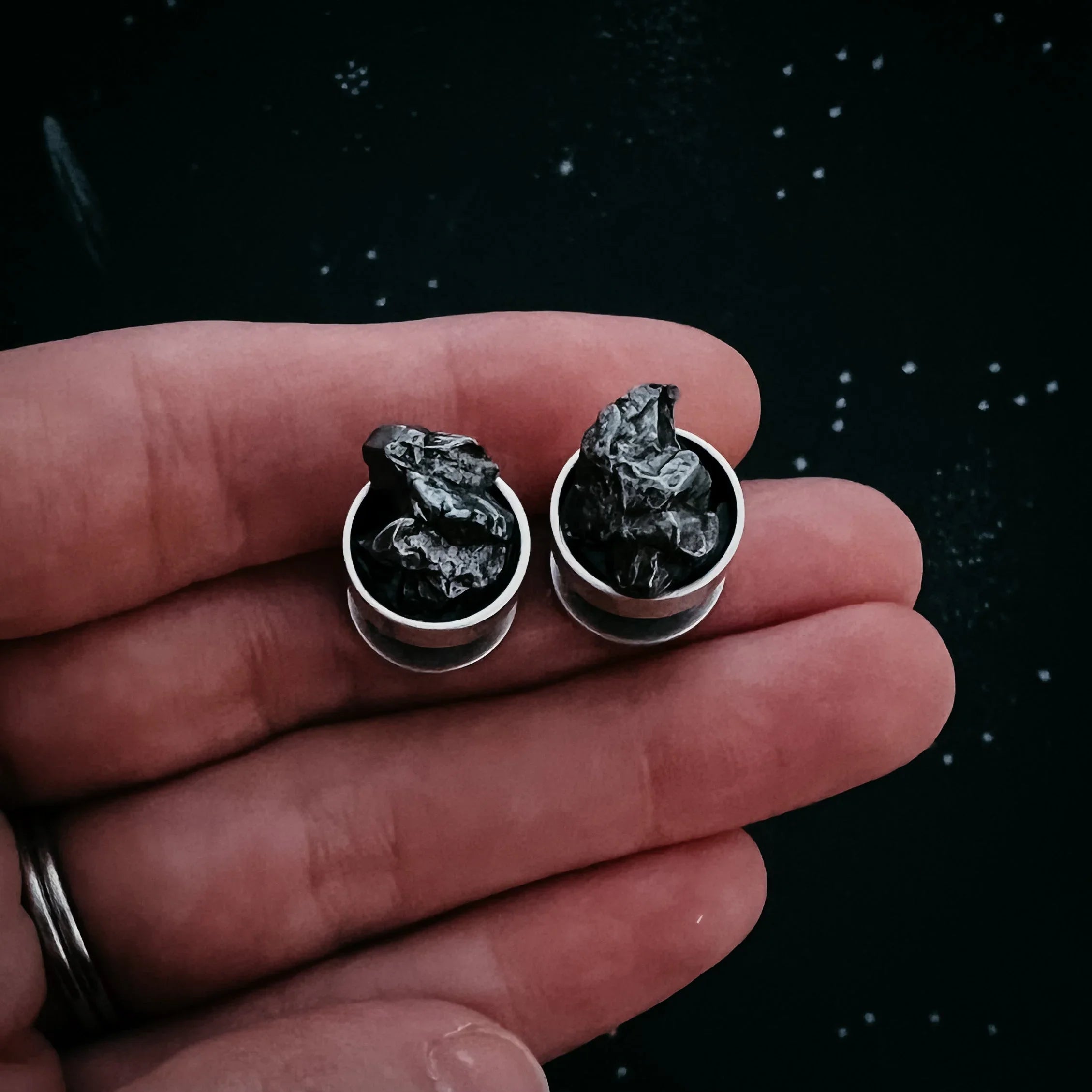 Hand holding pair of silver stud earrings with black textured stones against dark background