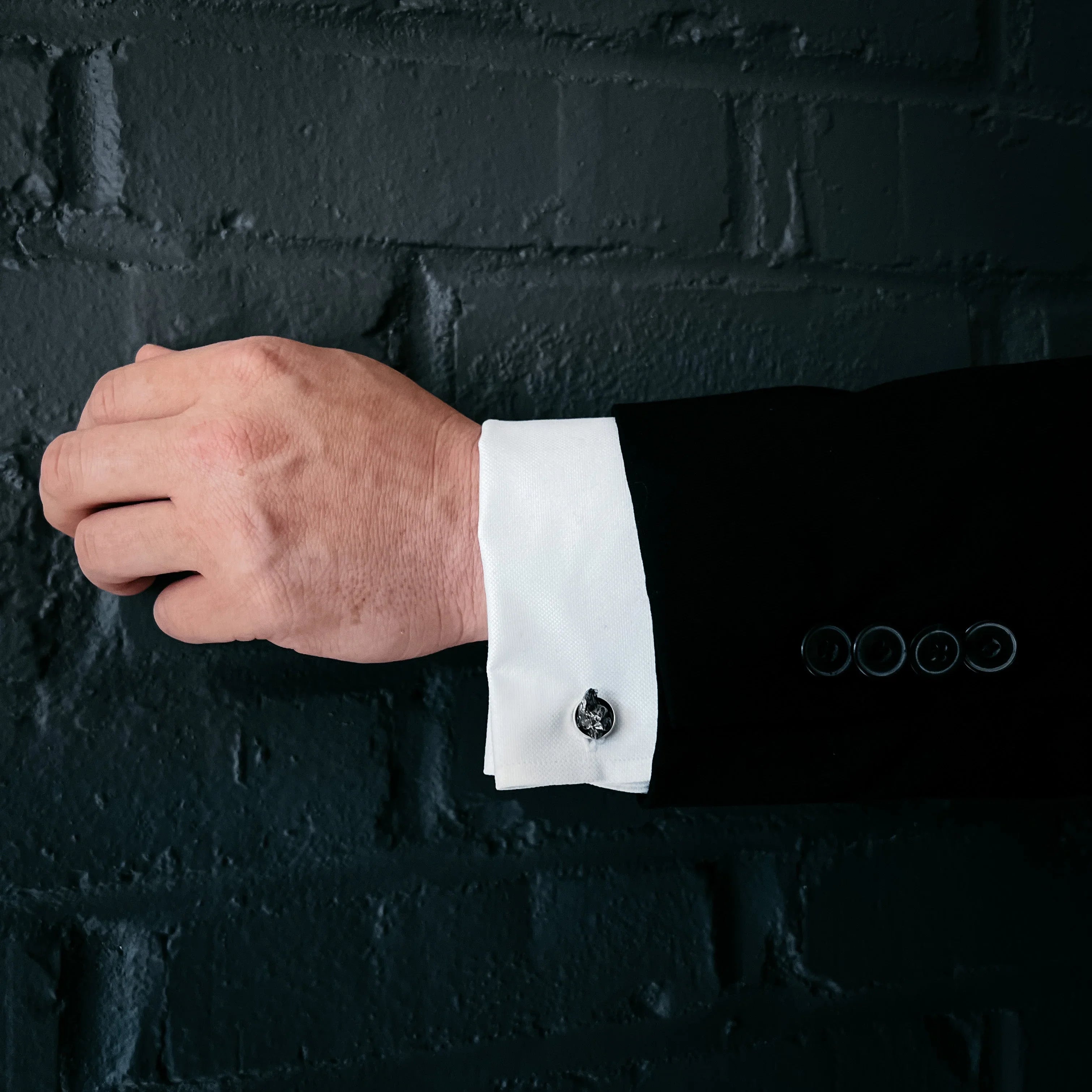 Close-up of a man's hand wearing a black suit with white cuff and silver cufflink against a dark brick wall