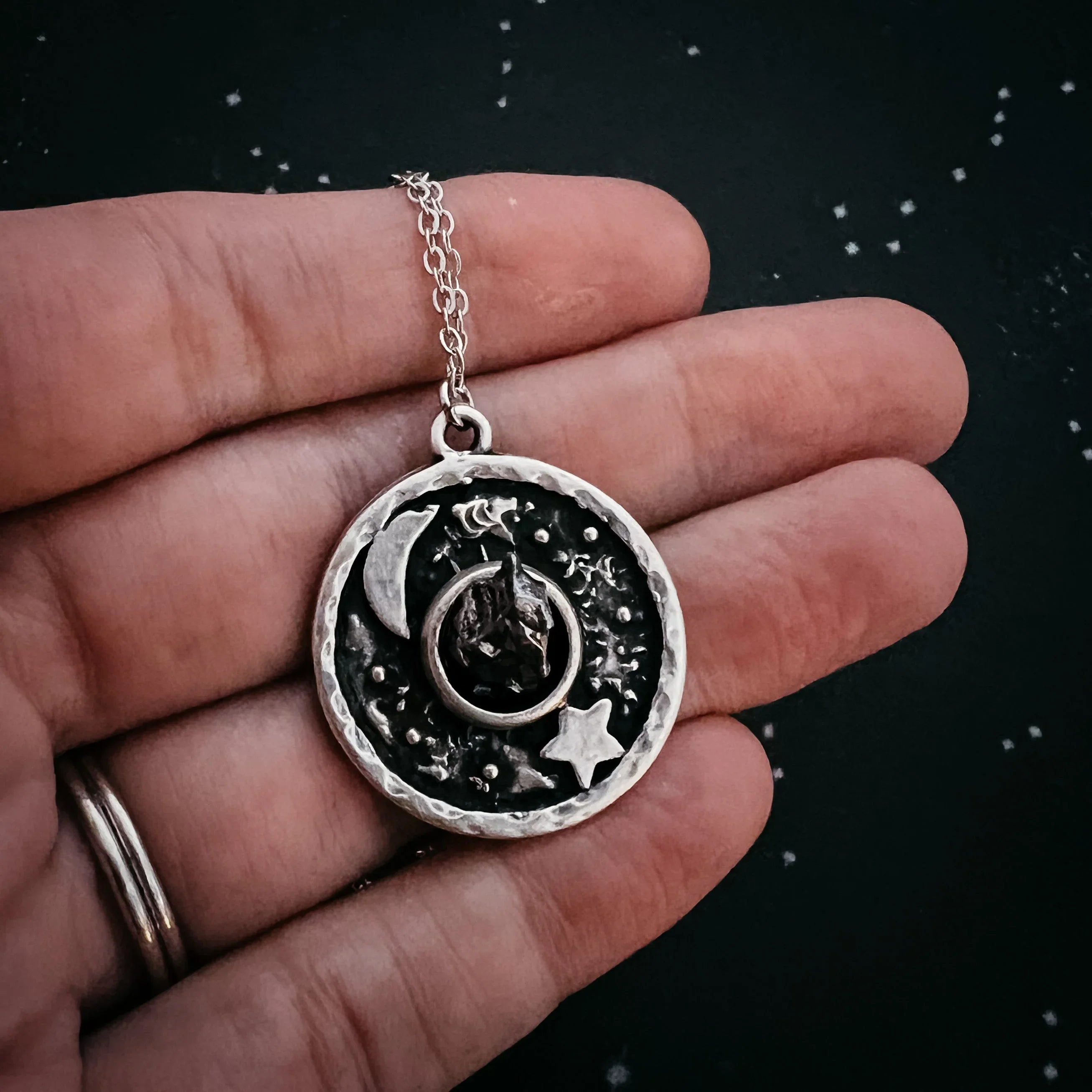 Hand holding a silver pendant necklace with moon, star, and wolf head design on black background
