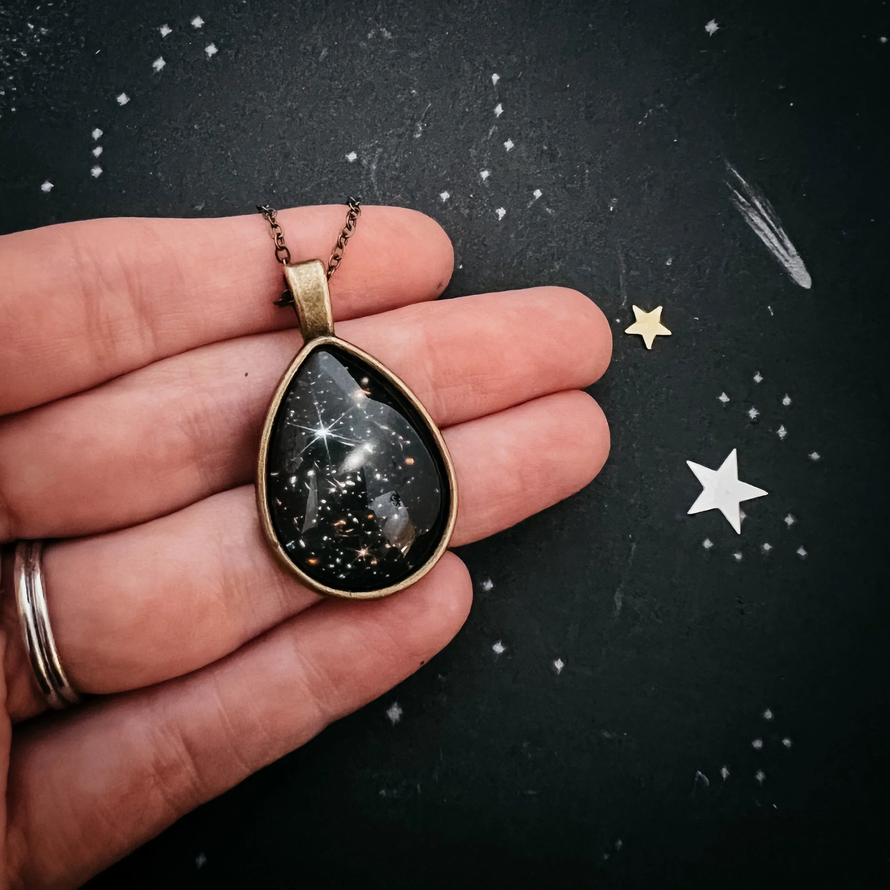 Hand holding cosmic galaxy teardrop pendant necklace on dark starry background, Qu!rk Boutique gift
