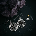 Cosmic-themed silver earrings with moon and star design, displayed on a dark starry background with crystal cluster, boutique jewelry by Qu!rk.