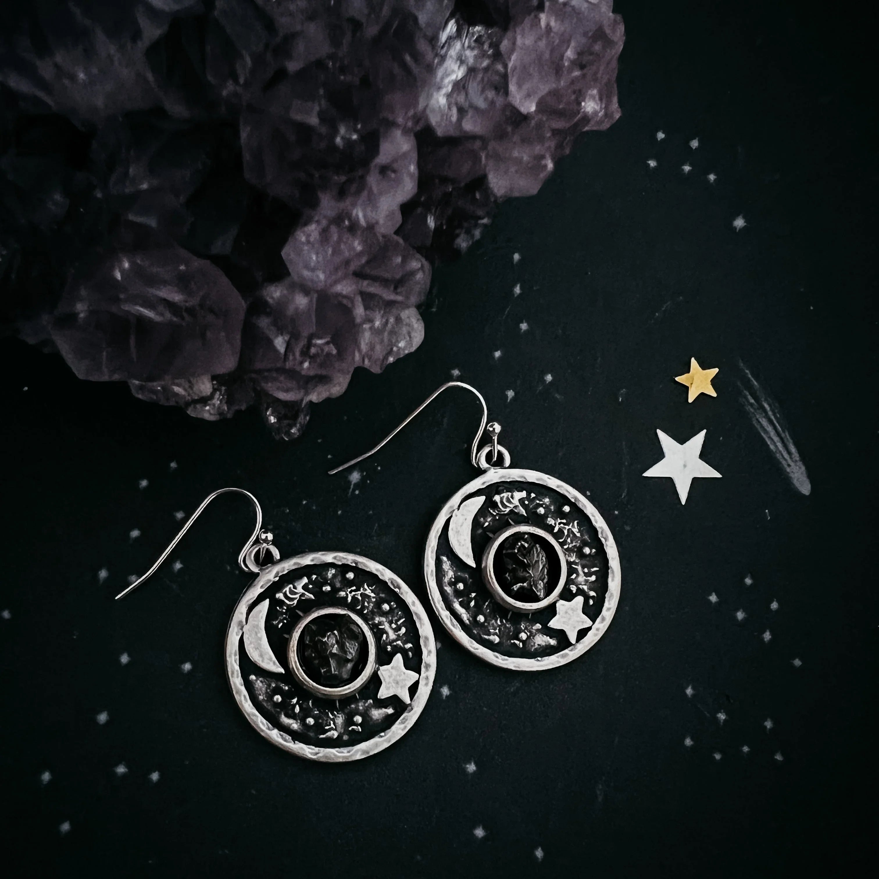 Cosmic-themed silver earrings with moon and star design, displayed on a dark starry background with crystal cluster, boutique jewelry by Qu!rk.