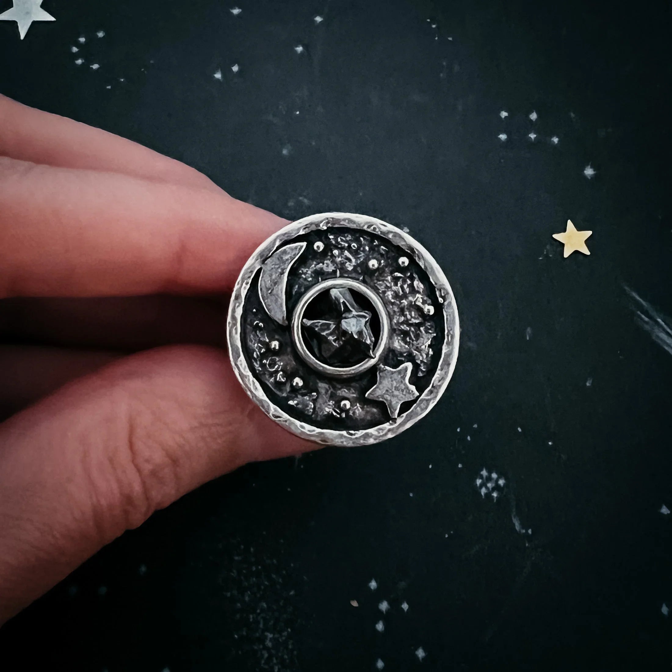 Hand holding round silver celestial ring with moon, stars, and textured black background