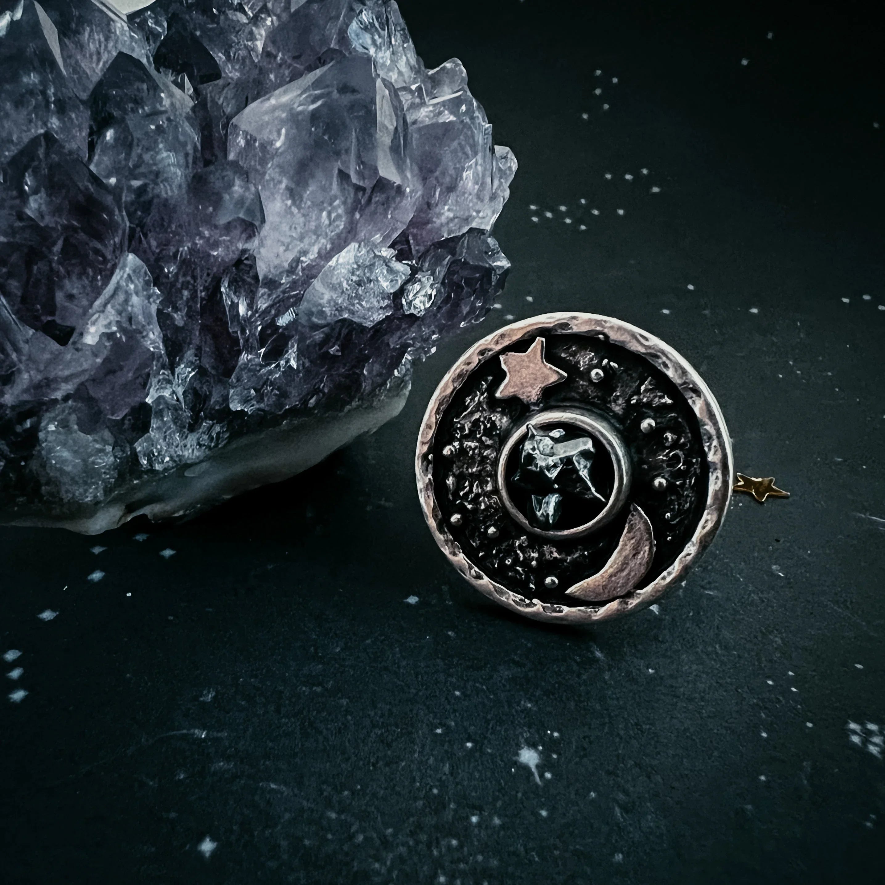Round cosmic-themed ring with star, moon, and crystal design beside purple amethyst crystal