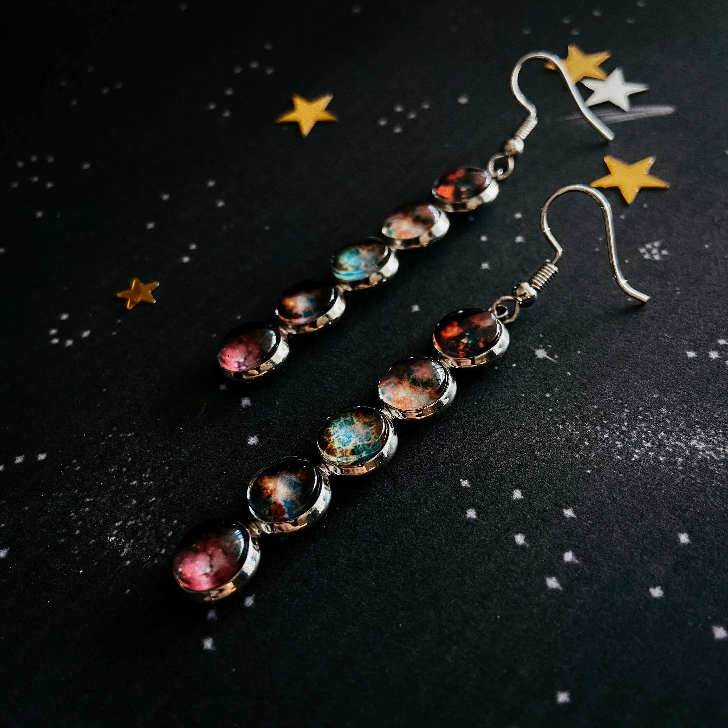 Long dangling silver earrings with multicolored galaxy-inspired stones on starry black background