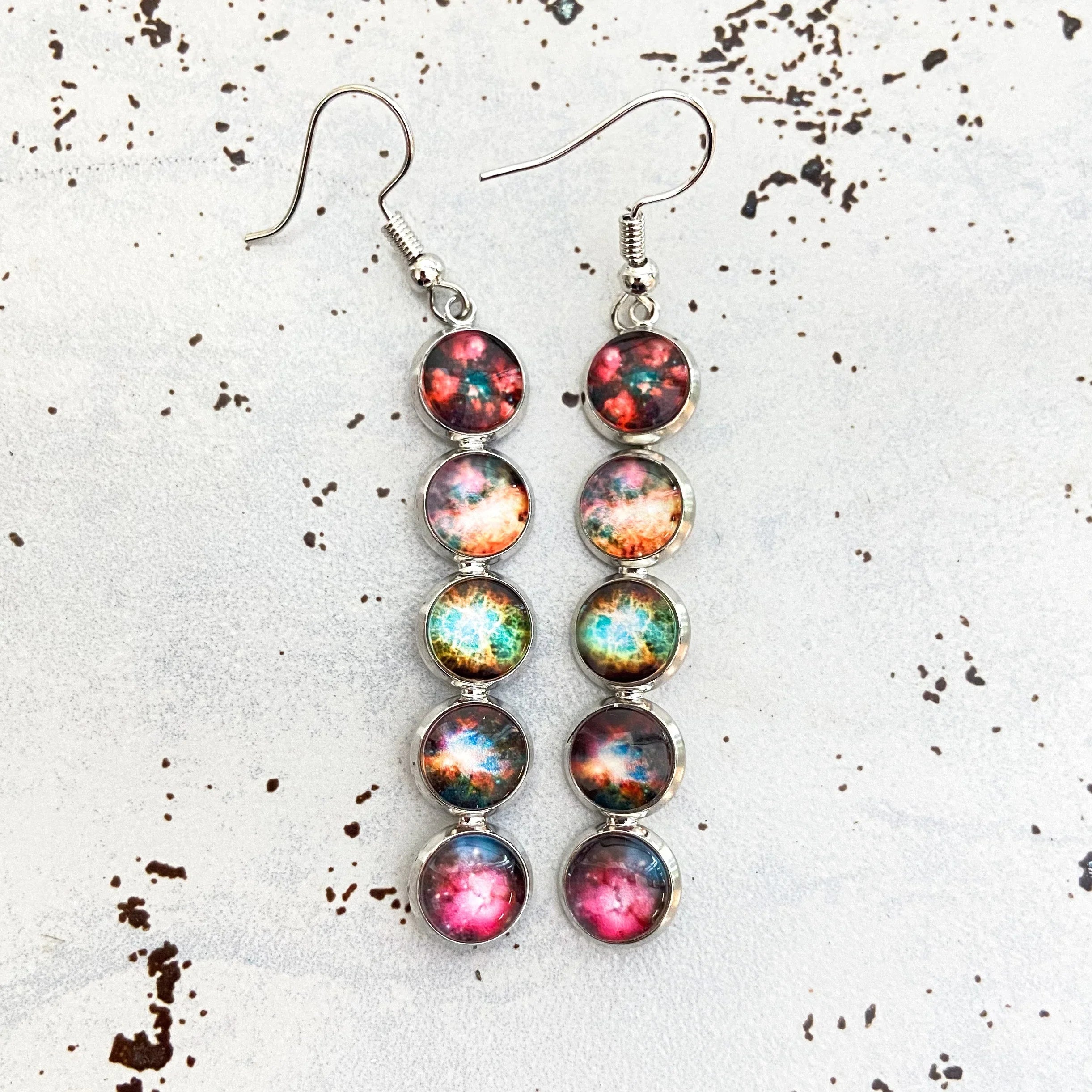 Long silver dangle earrings with four multicolored galaxy-themed round charms on a white speckled background