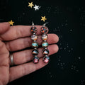 Hand holding long galaxy-themed earrings with colorful space designs on black starry background
