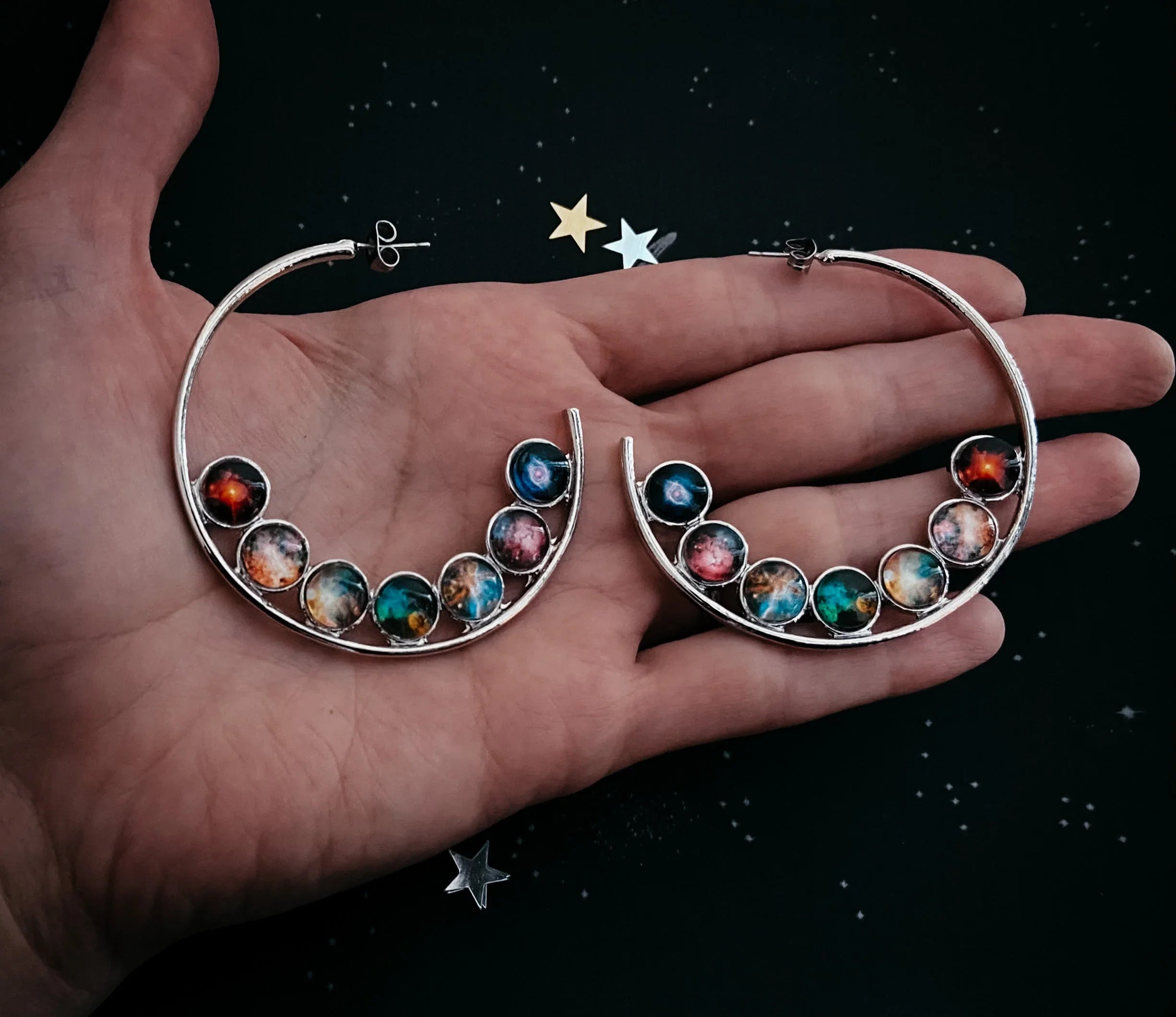 Hand holding silver hoop earrings with colorful galaxy-themed round stones on a black starry background