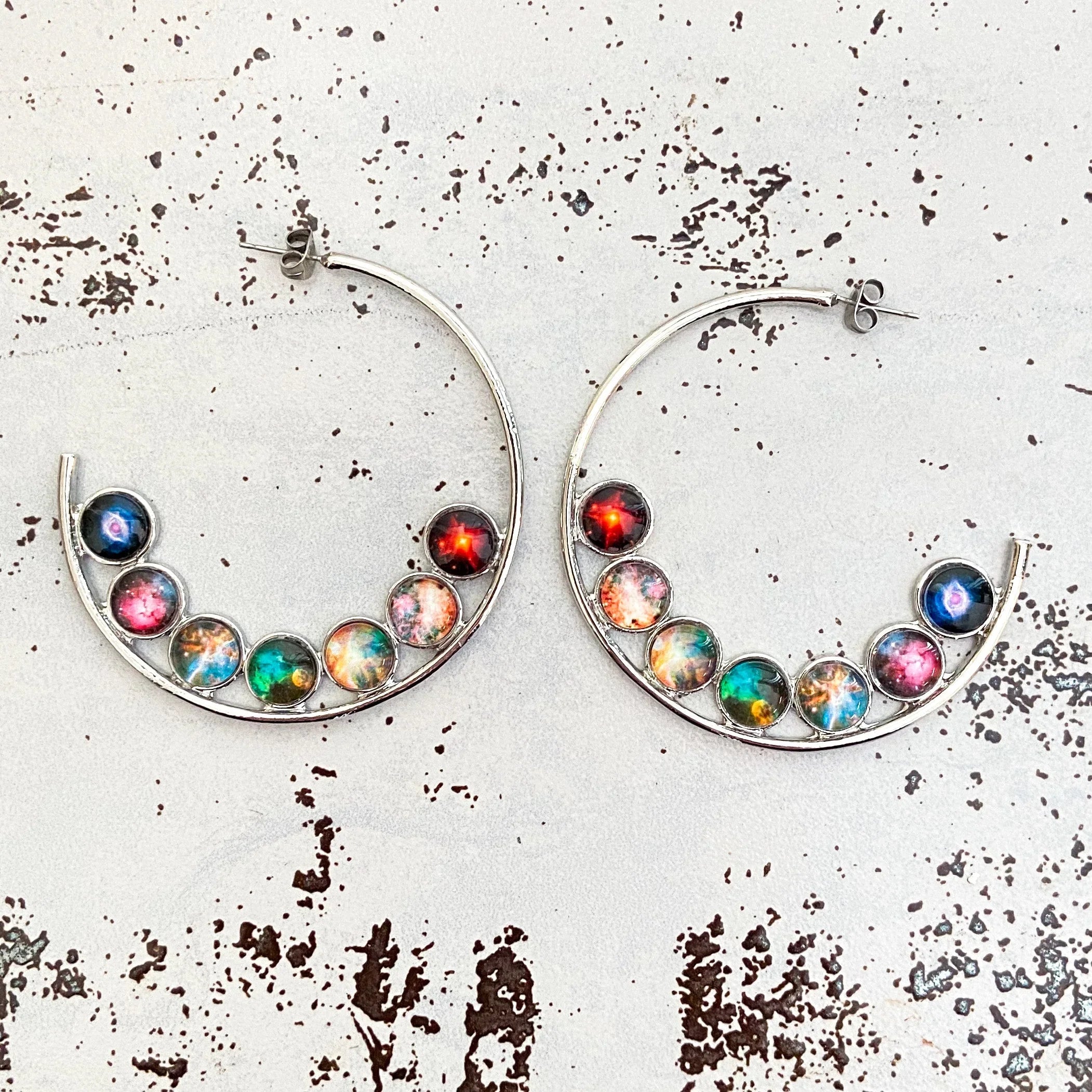 Silver hoop earrings with colorful galaxy-themed round charms on textured white background
