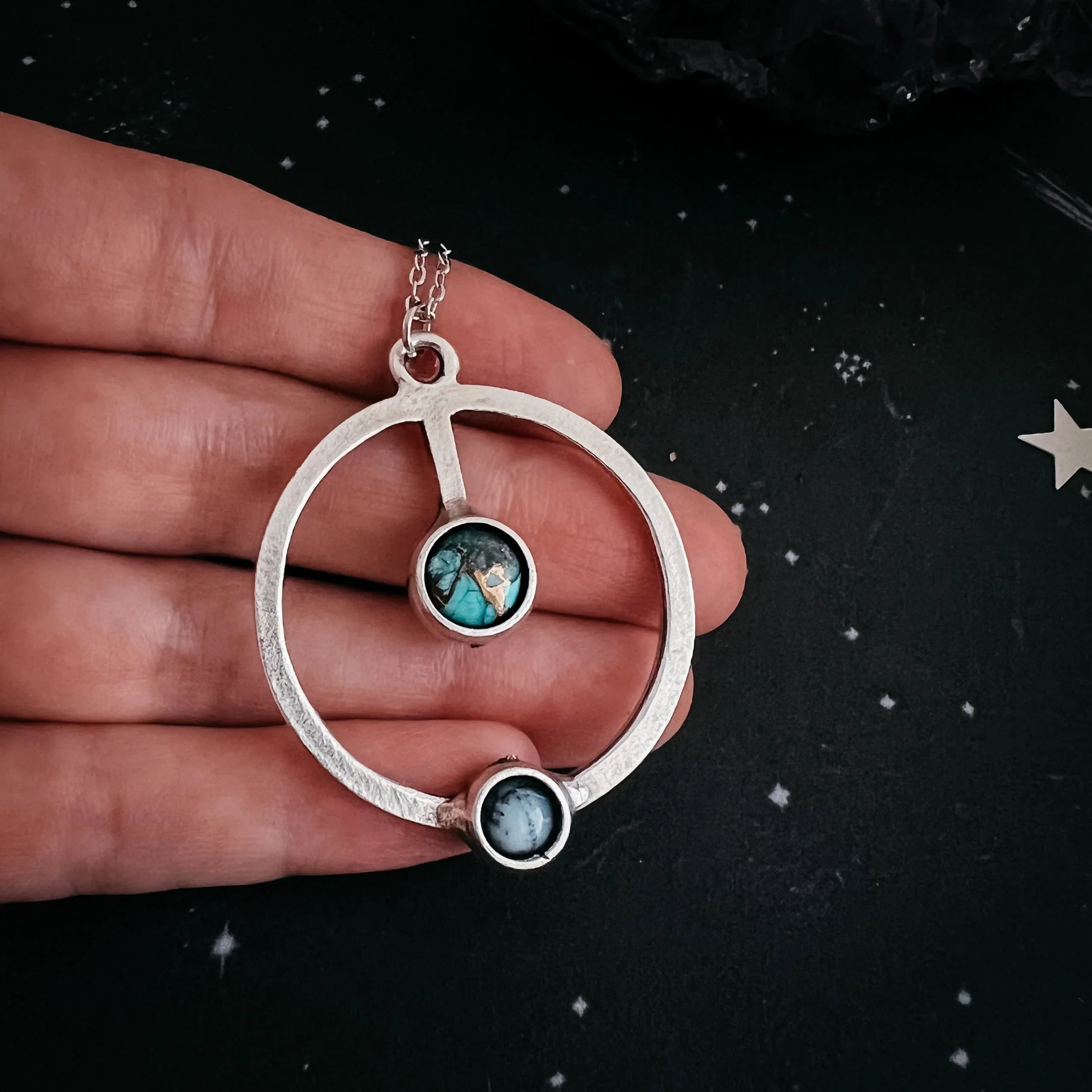 Hand holding silver circular pendant necklace with two round gemstone accents on starry black background
