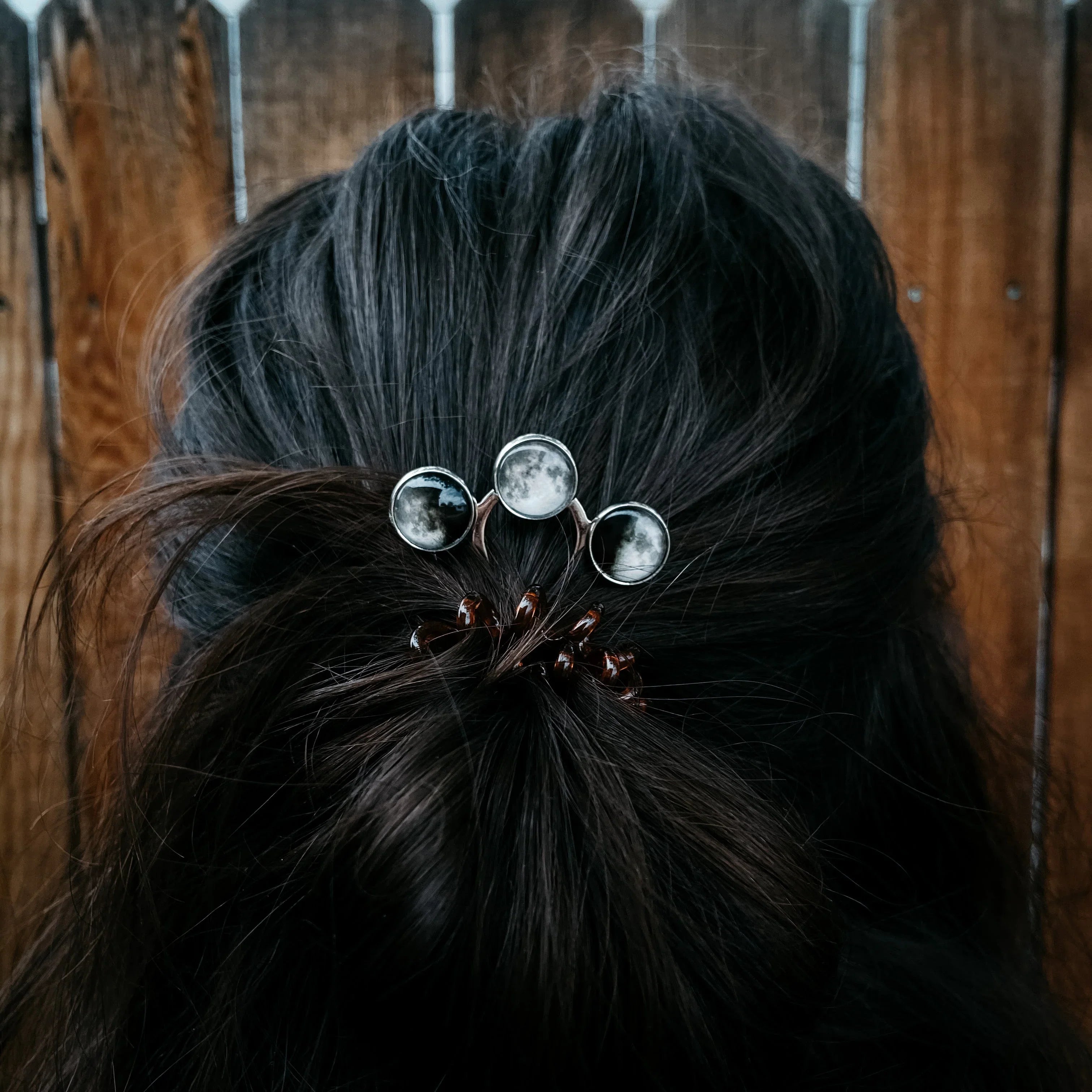 Dark hair half up secured with a hair clip featuring moon phase designs against a wooden fence background
