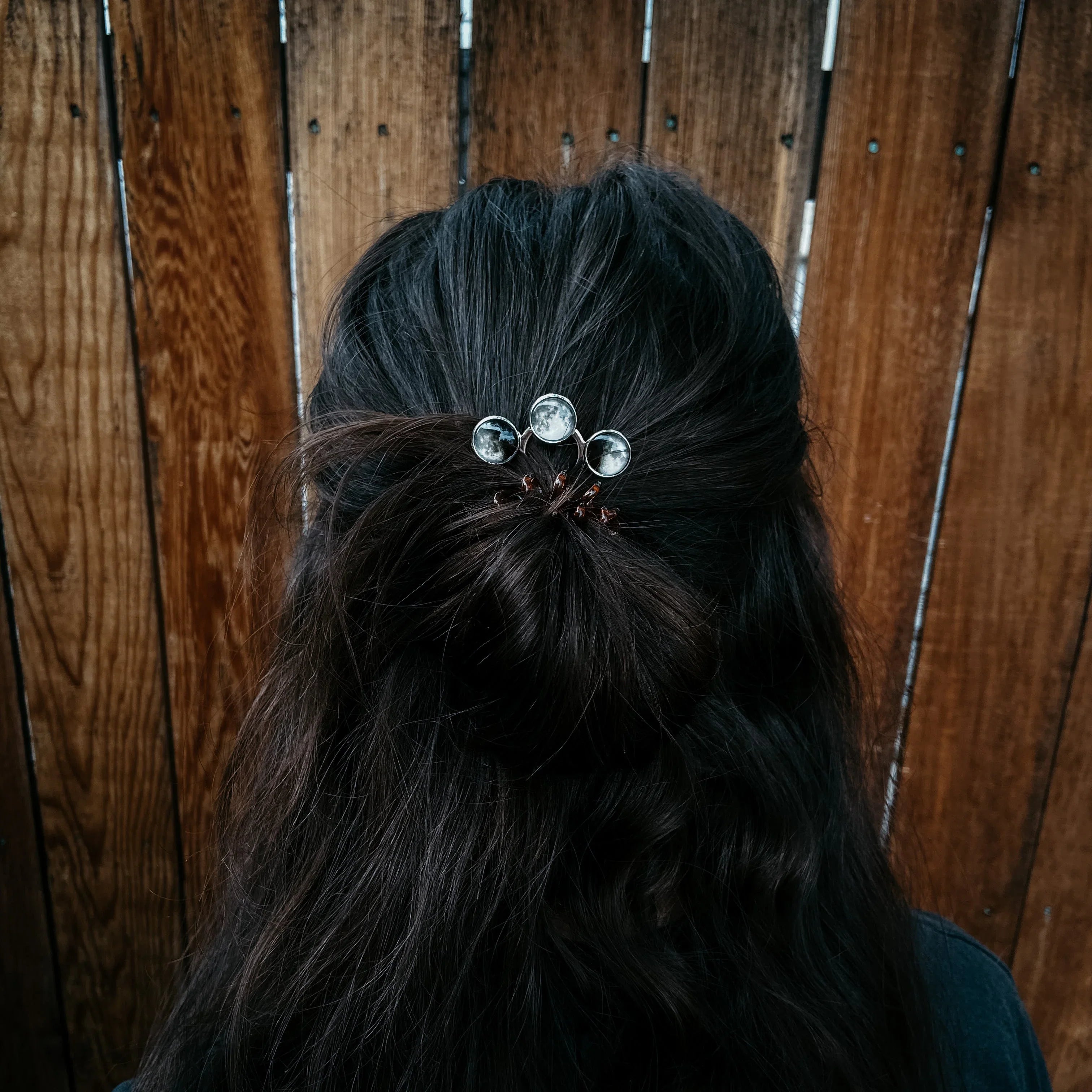 Dark brown hair styled in a bun with a silver hairpin featuring three moon phase designs against a wooden fence background