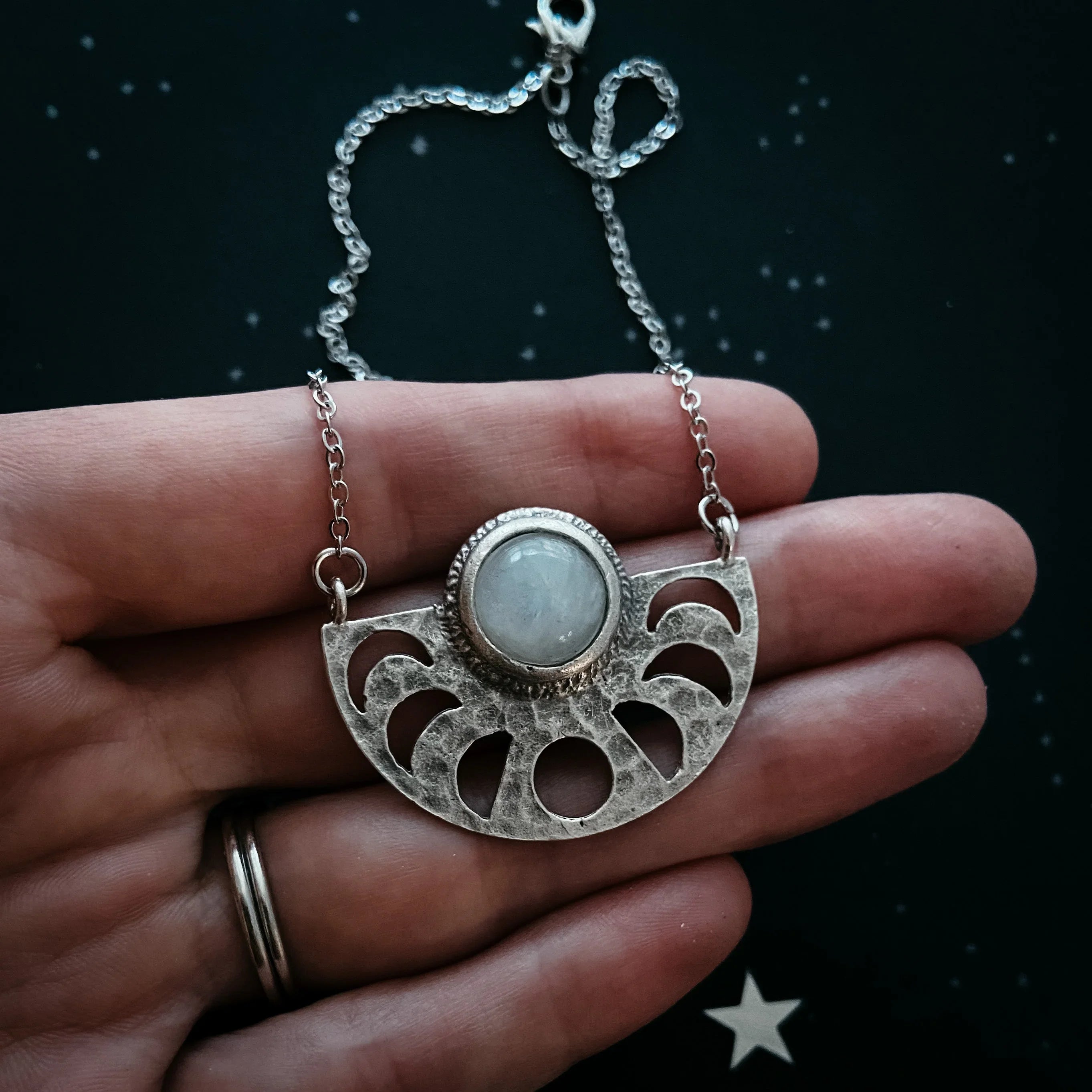 Hand holding silver moon phase necklace with round white gemstone on black starry background