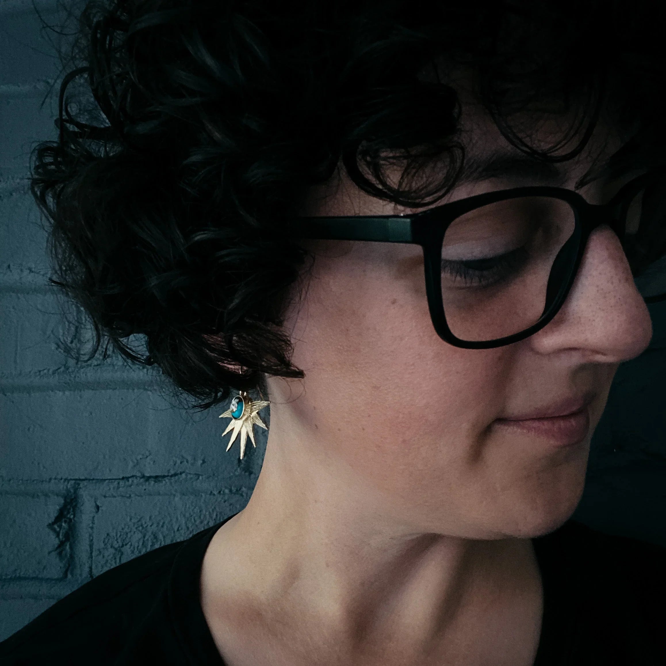 Close-up of woman with curly hair wearing black glasses and gold sunburst turquoise earrings