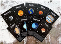 Set of enamel pins depicting planets Mercury, Venus, Earth, Mars, Jupiter, Saturn, Uranus, Neptune, and Pluto on black starry cards