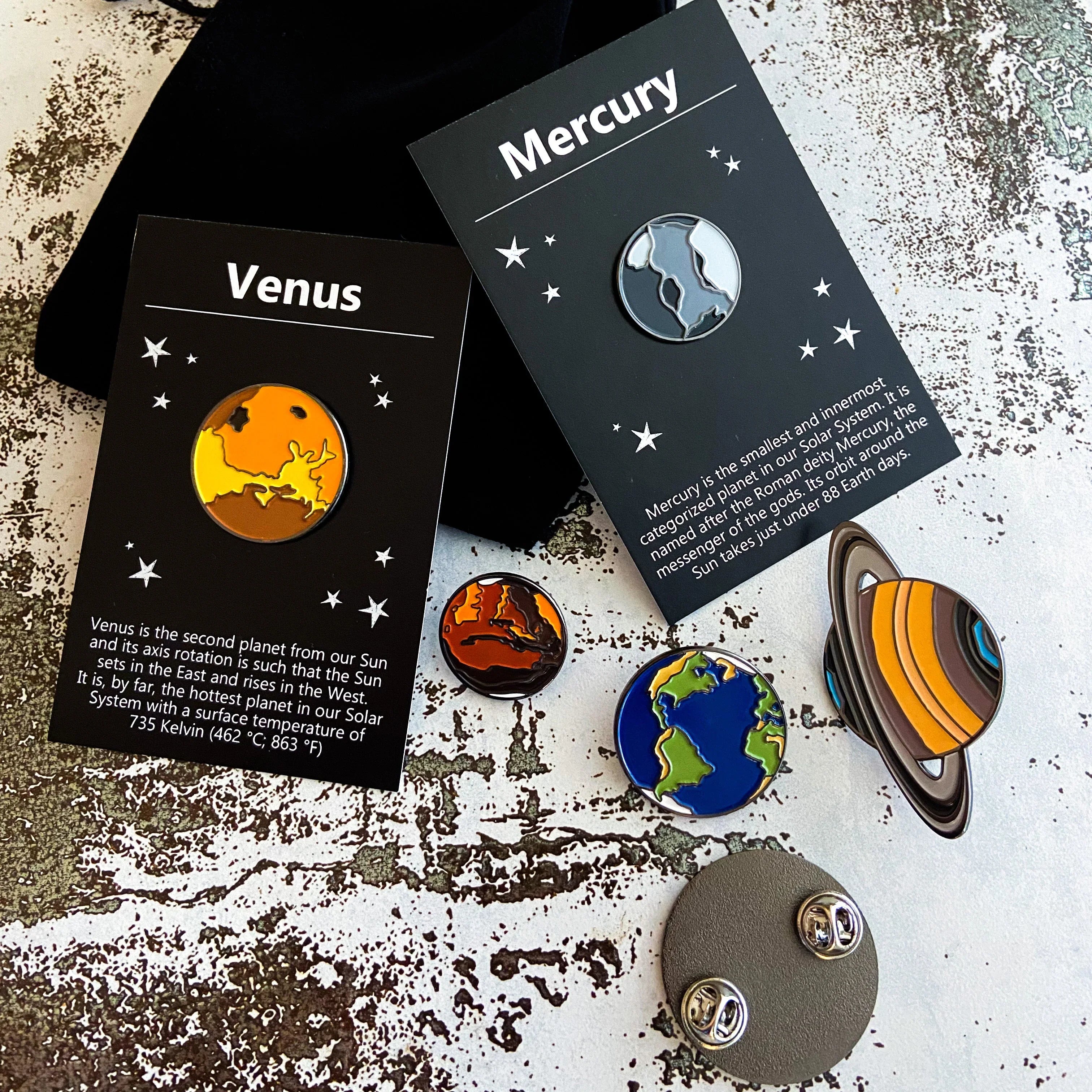 Enamel pins of planets Mercury and Venus on black cards with planet facts, plus Earth, Saturn, and Mars pins