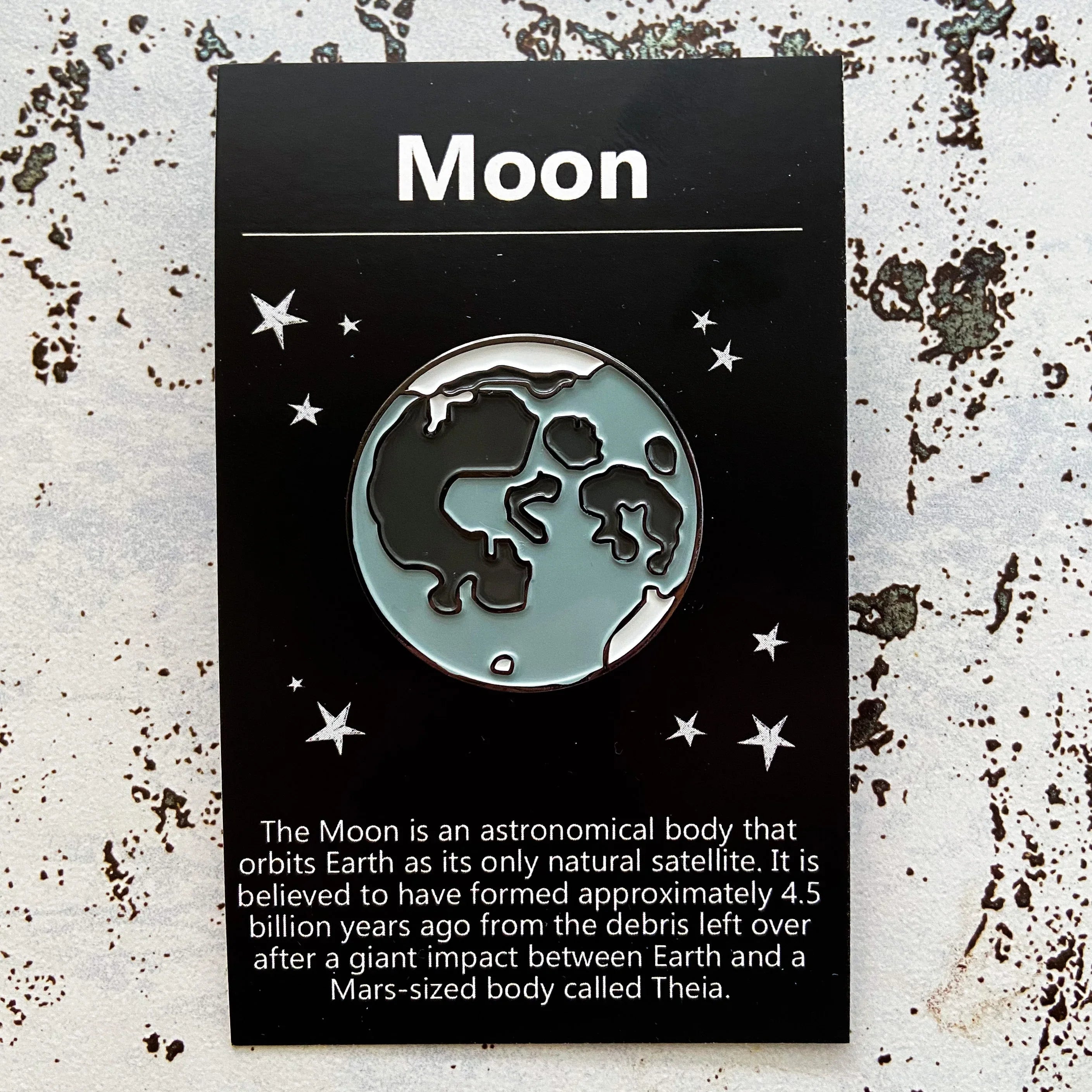 Moon enamel pin on black card with stars and text about moon formation and orbiting Earth