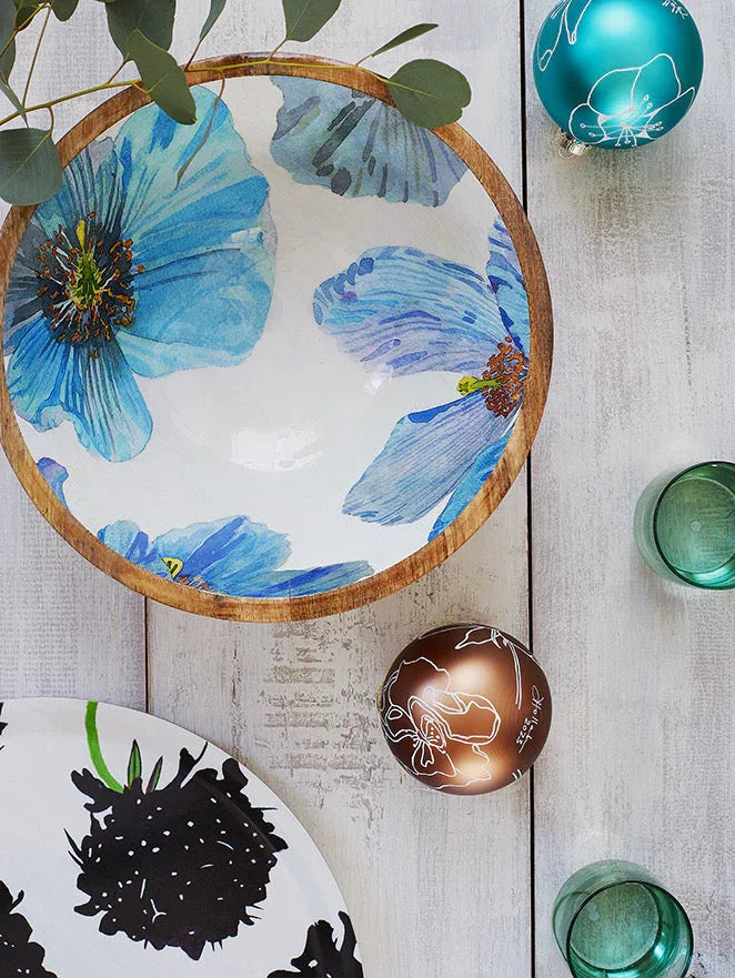Wooden bowl with blue floral design, decorative Christmas baubles, and green glass cups on white wooden table