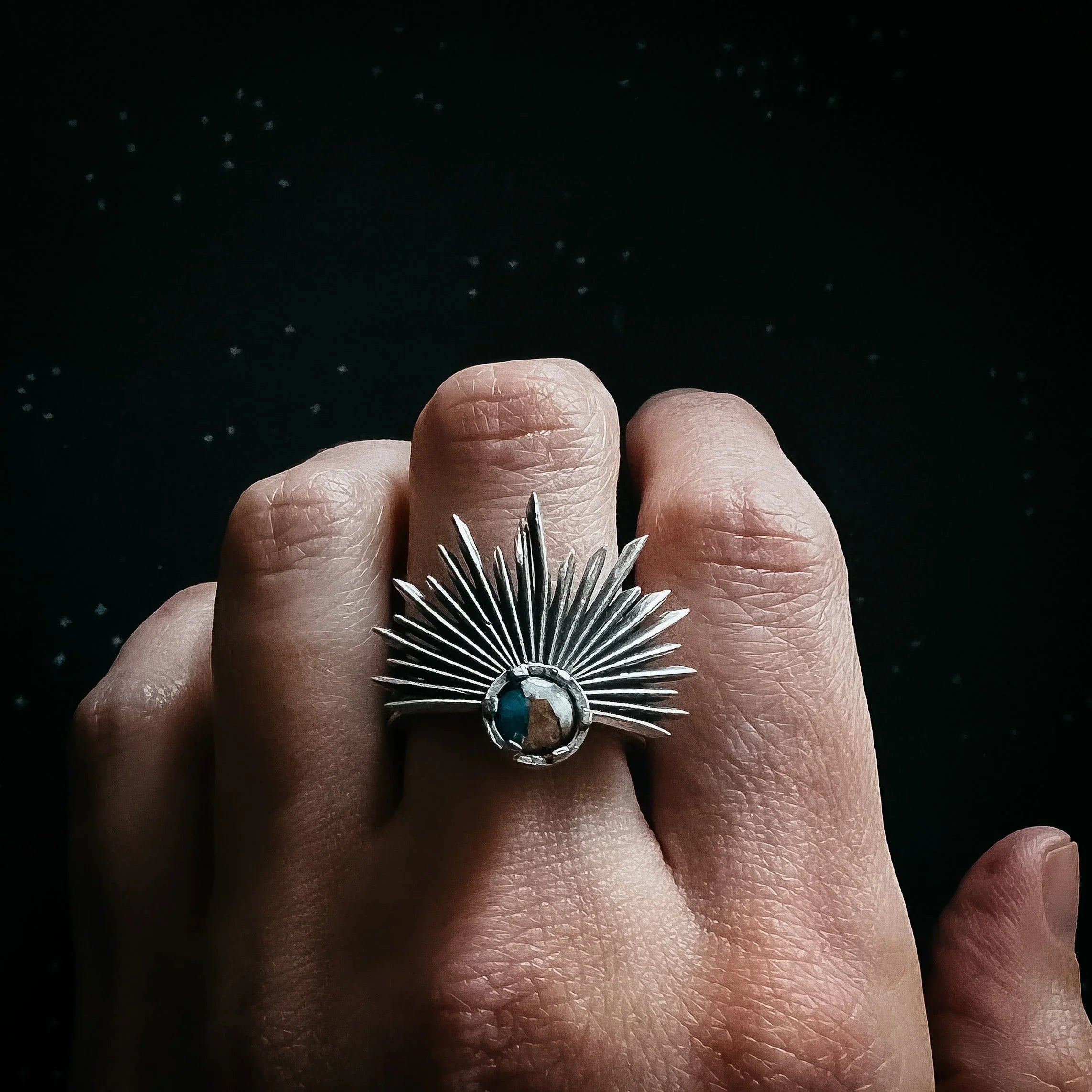 Close-up of hand wearing silver ring with sunburst design and central round stone on dark background
