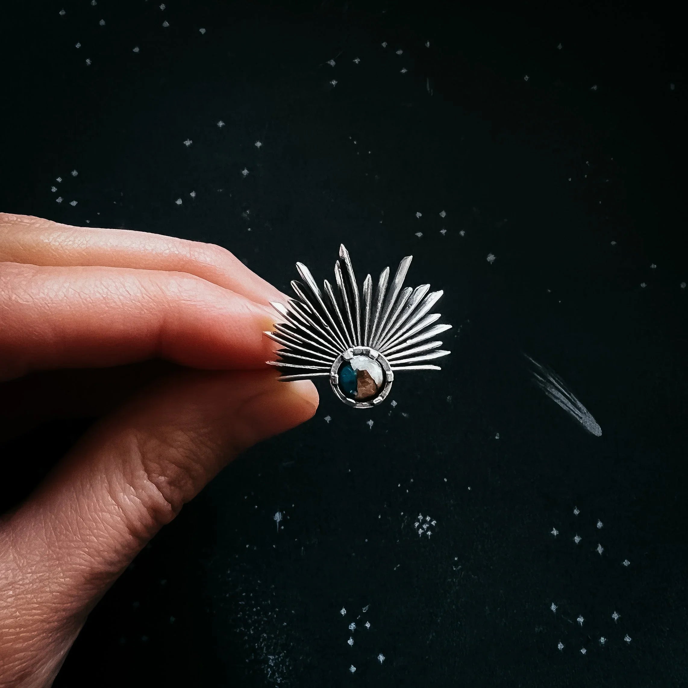 Close-up of hand holding silver sunburst ring with blue and brown stone on black background