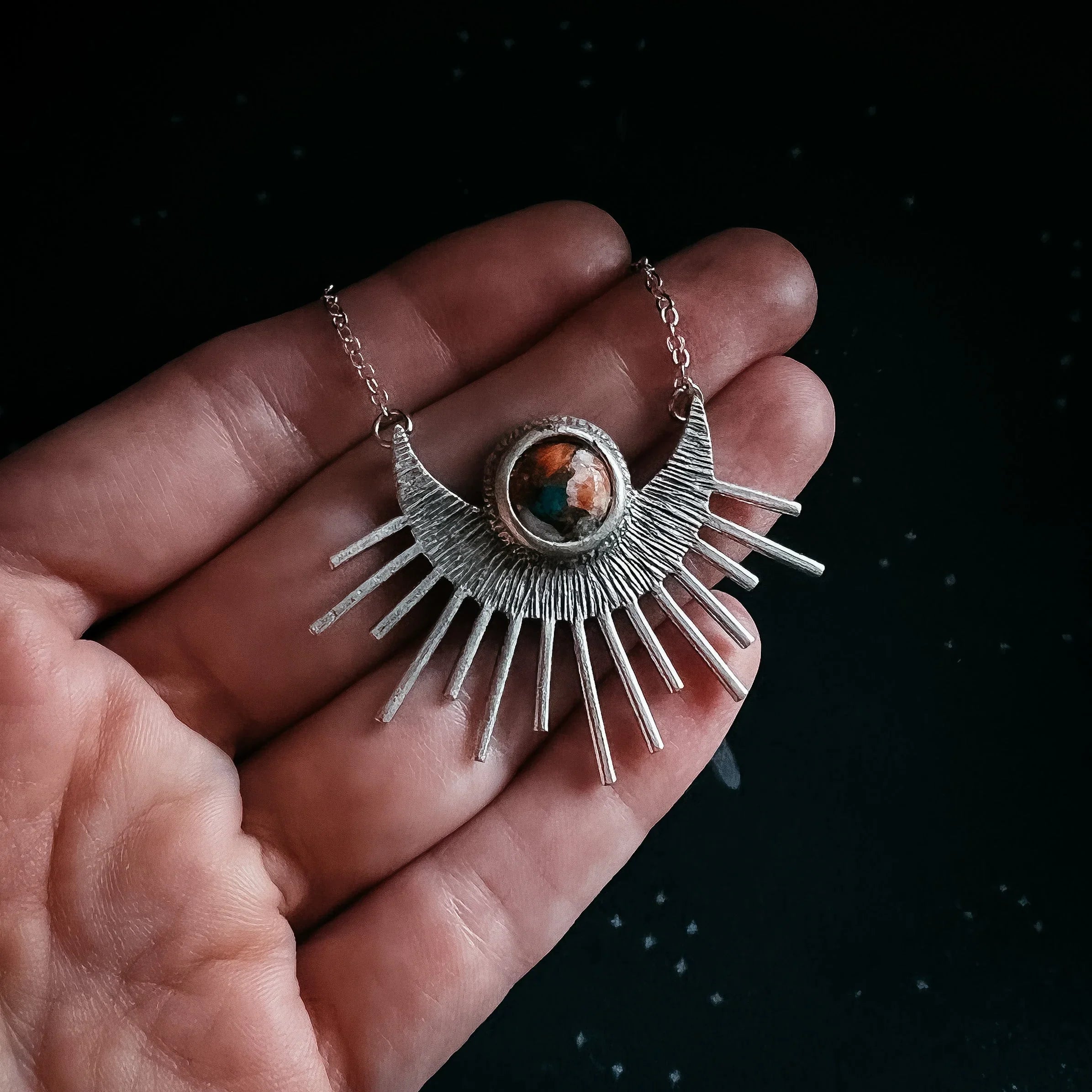 Hand holding silver sunburst necklace with colorful stone pendant on black background