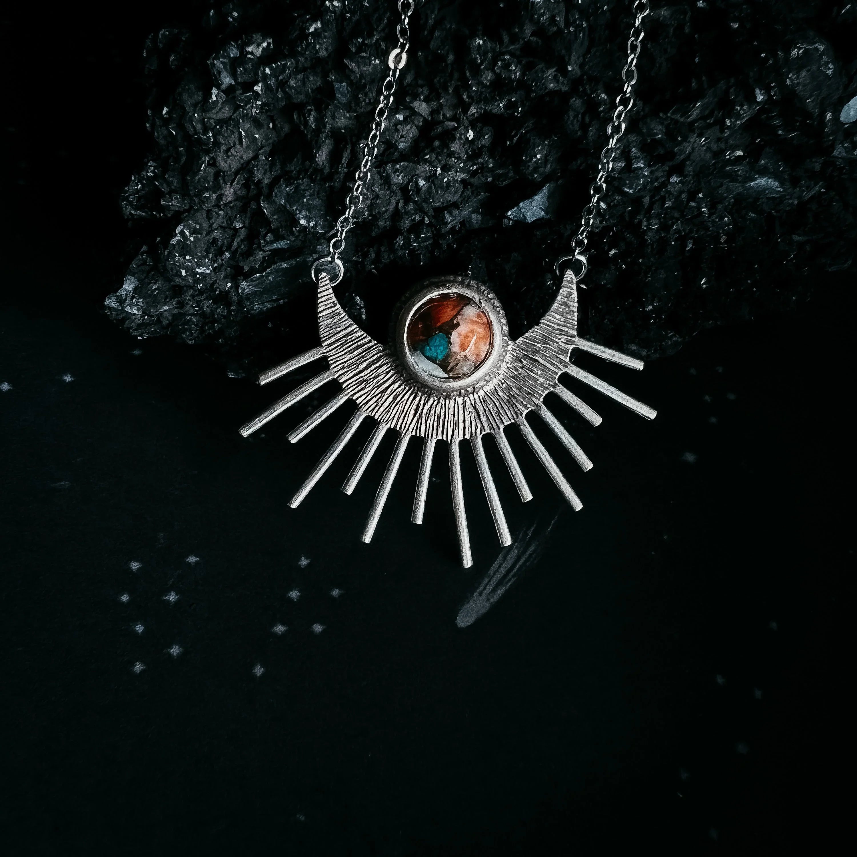 Silver sunburst pendant necklace with multicolored stone centerpiece on black textured background