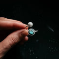 Hand holding an open silver ring with round white and turquoise stones against a black background