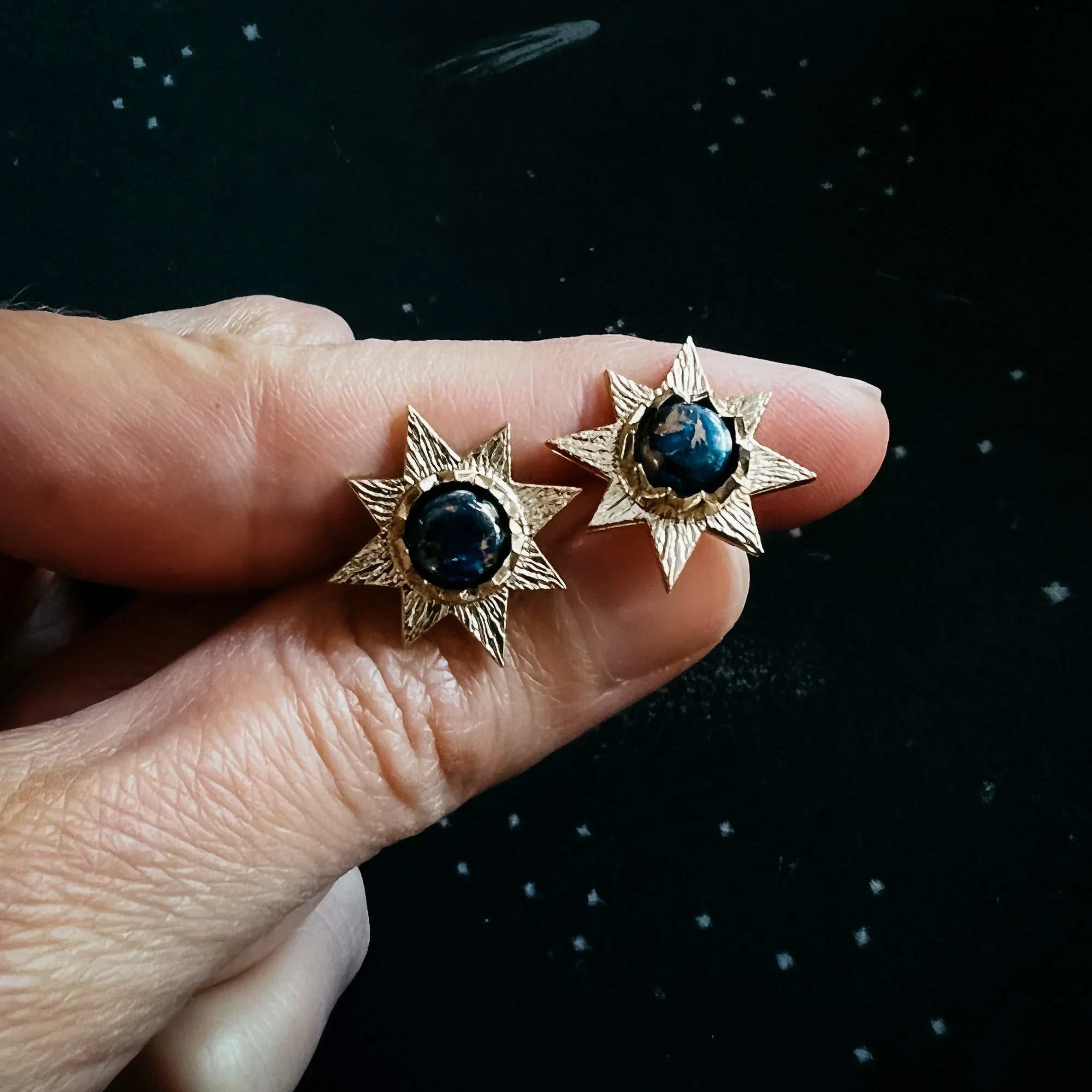 Hand holds gold starburst earrings with dark blue stones, cosmic jewelry at Qu!rk Boutique