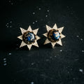 Gold starburst earrings with cosmic blue centers on a black speckled background at Qu!rk Boutique